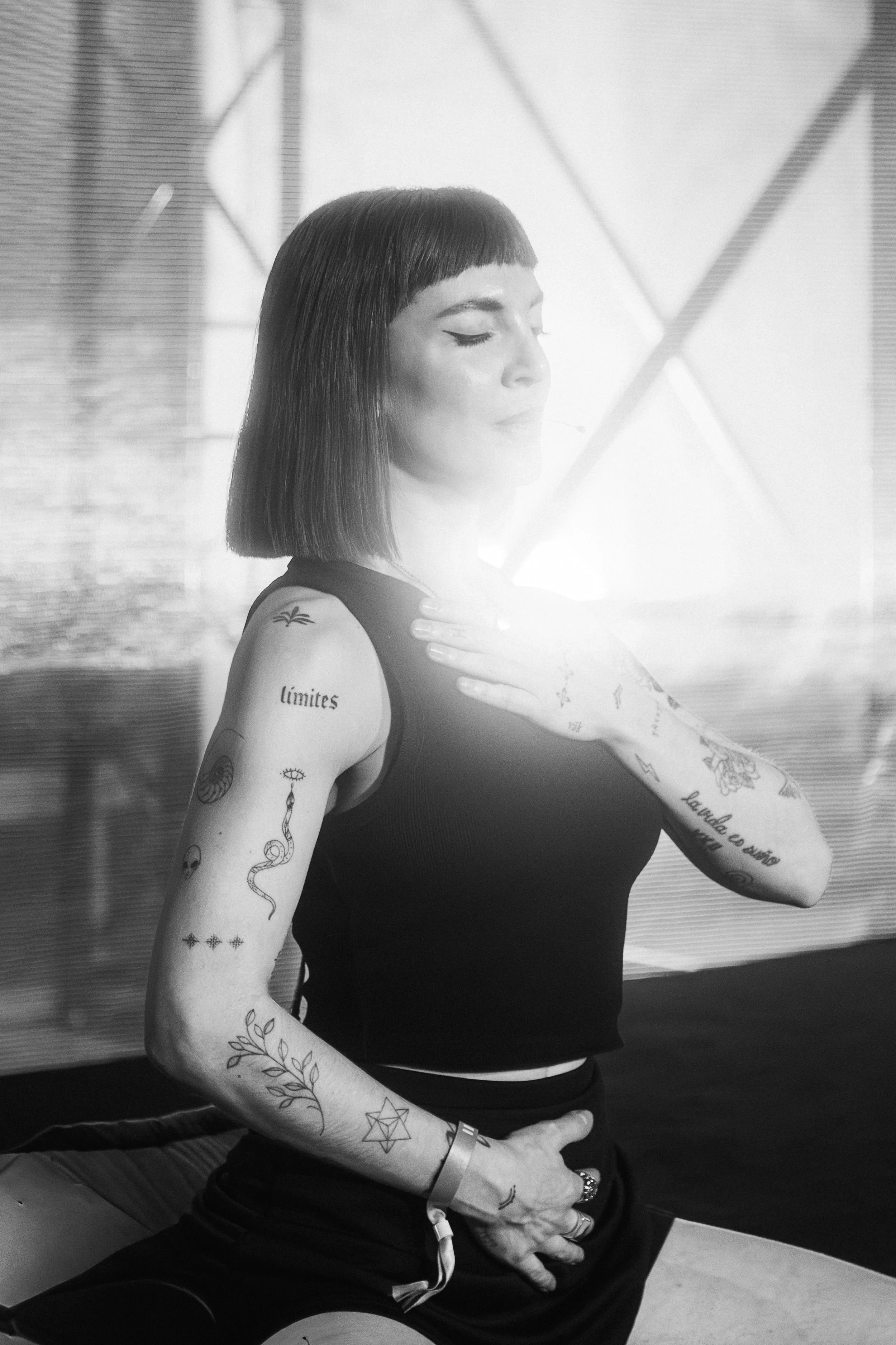 A woman with short black hair and multiple tattoos on her arms is in a stretch pose, wearing a black tank top and black pants. She is sitting against a wall with a grid pattern and appears to be in a gym or fitness studio.