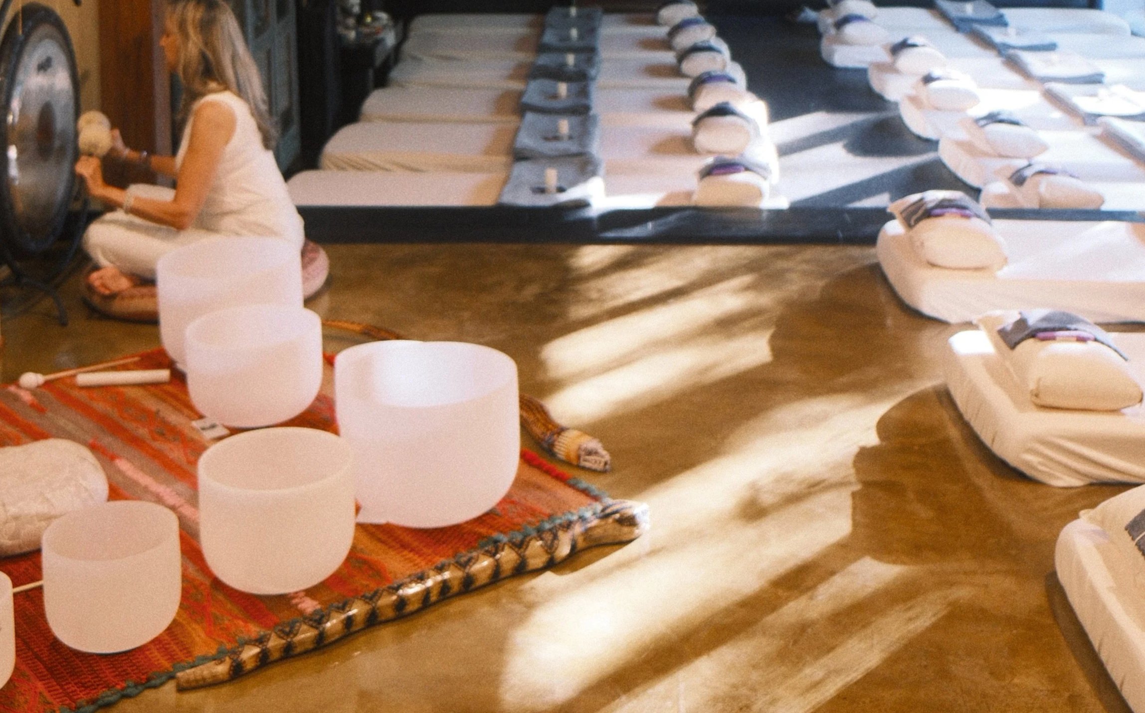 A woman is sitting cross-legged on a cushion in front of a large gong, playing a singing bowl with a mallet. Surrounding her are white crystal singing bowls and a colorful woven rug. There are chairs stacked in the background.