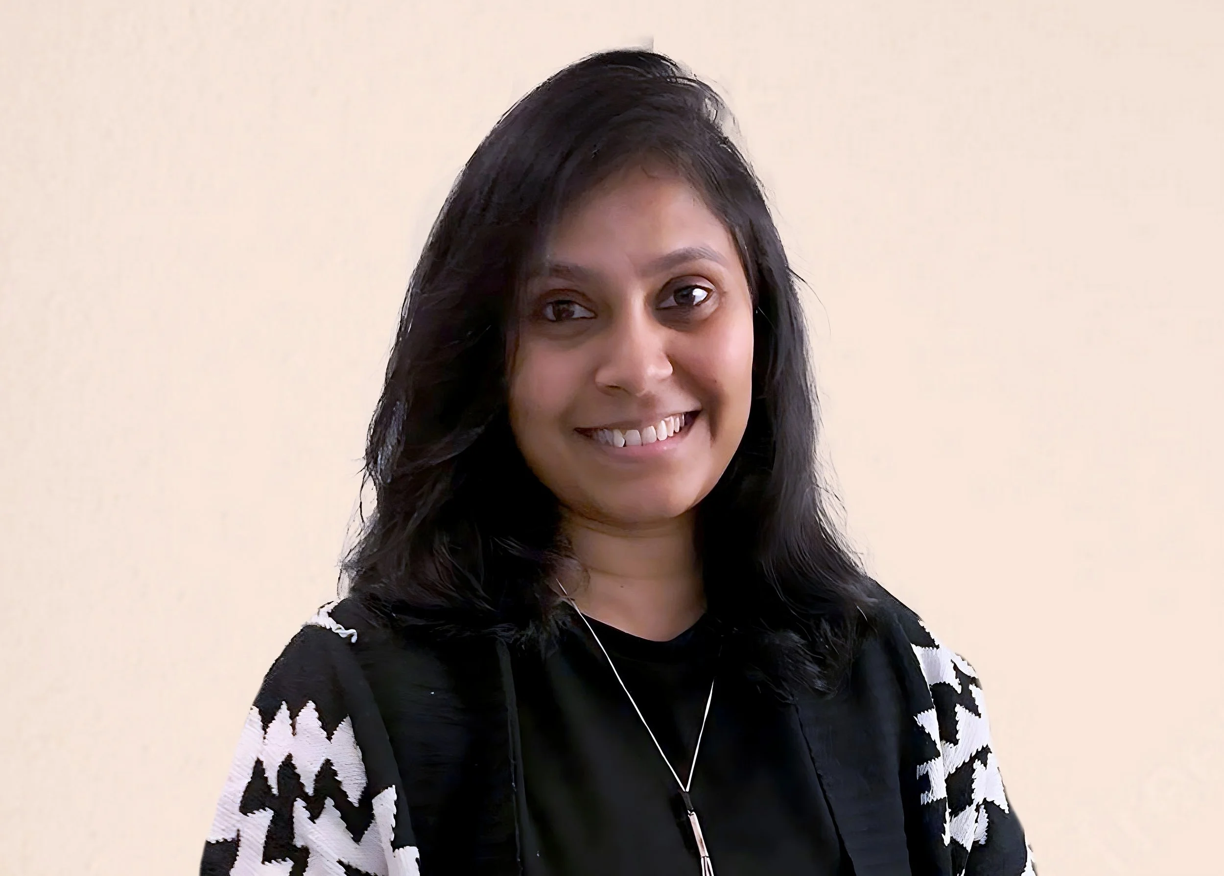 A woman with black hair smiling and wearing a black and white patterned top and a silver necklace against a plain beige background.