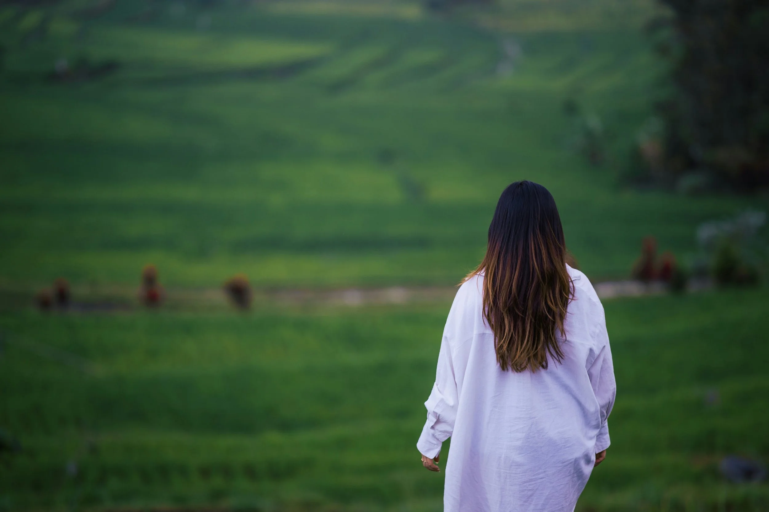 A woman with long dark hair, wearing a white shirt, is walking in a green grassy field with rolling hills in the background.