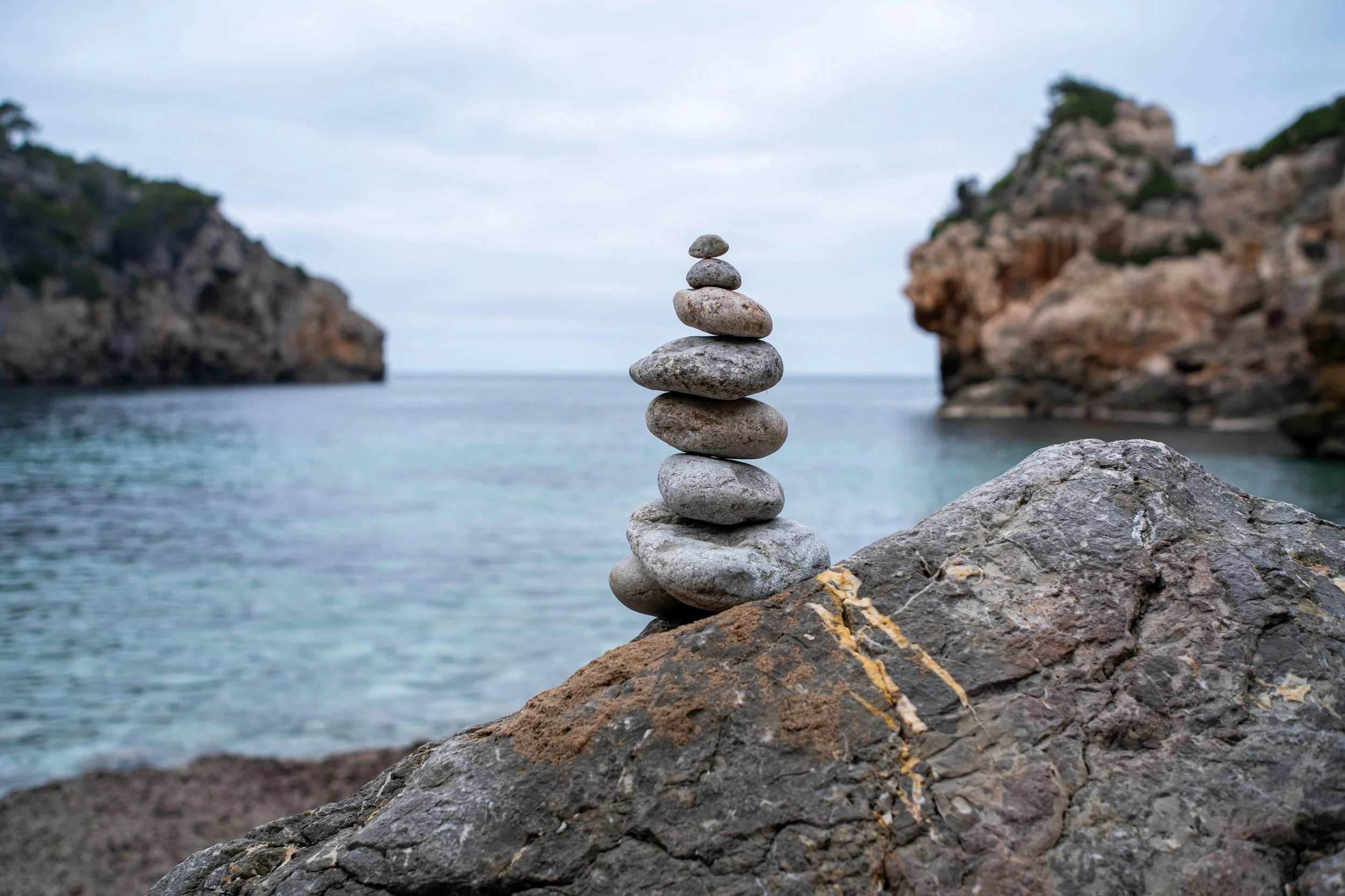 Stacked stones balanced on a large rock near the ocean with cliffs and water in background.