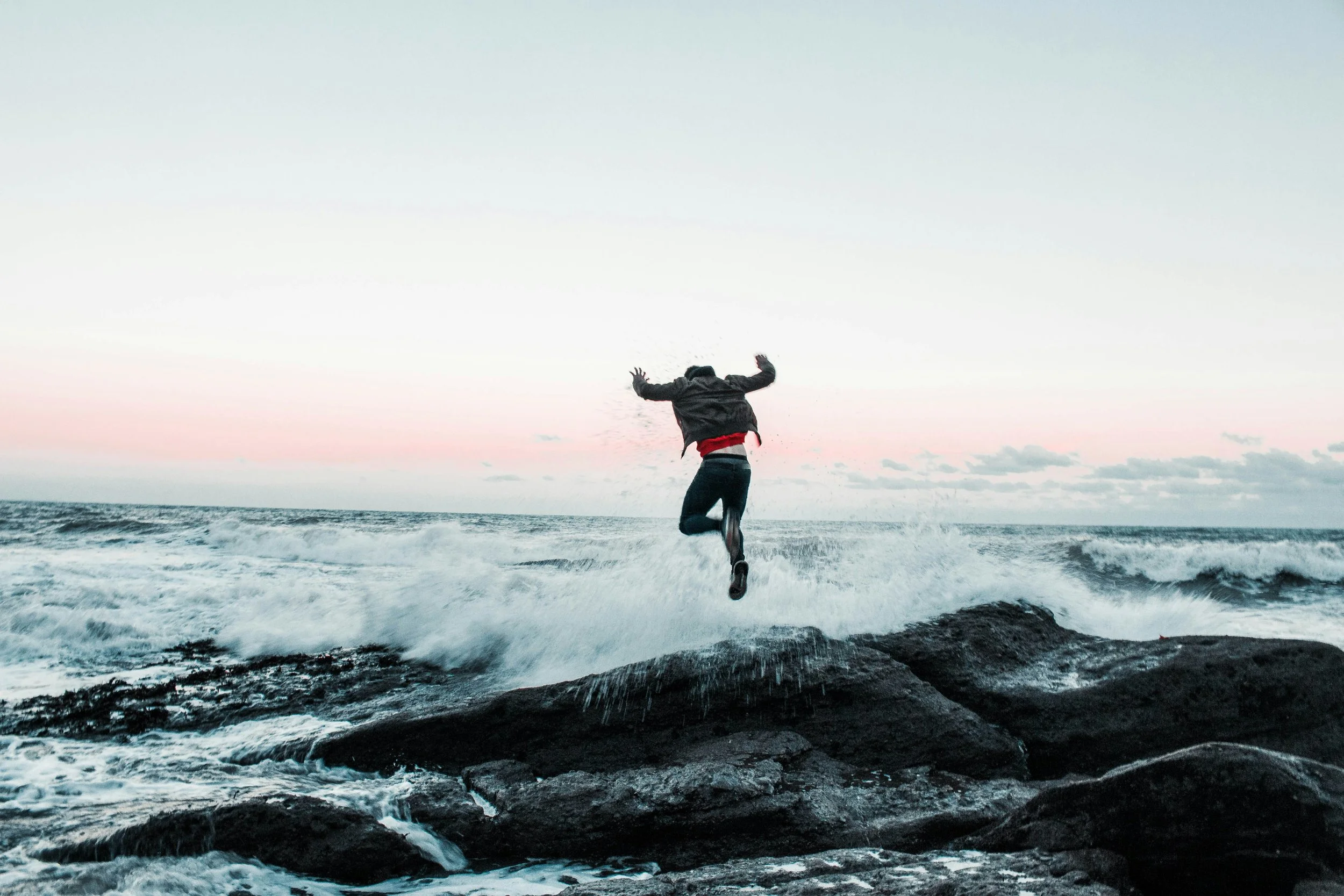 Person jumping between rocks near ocean during sunset