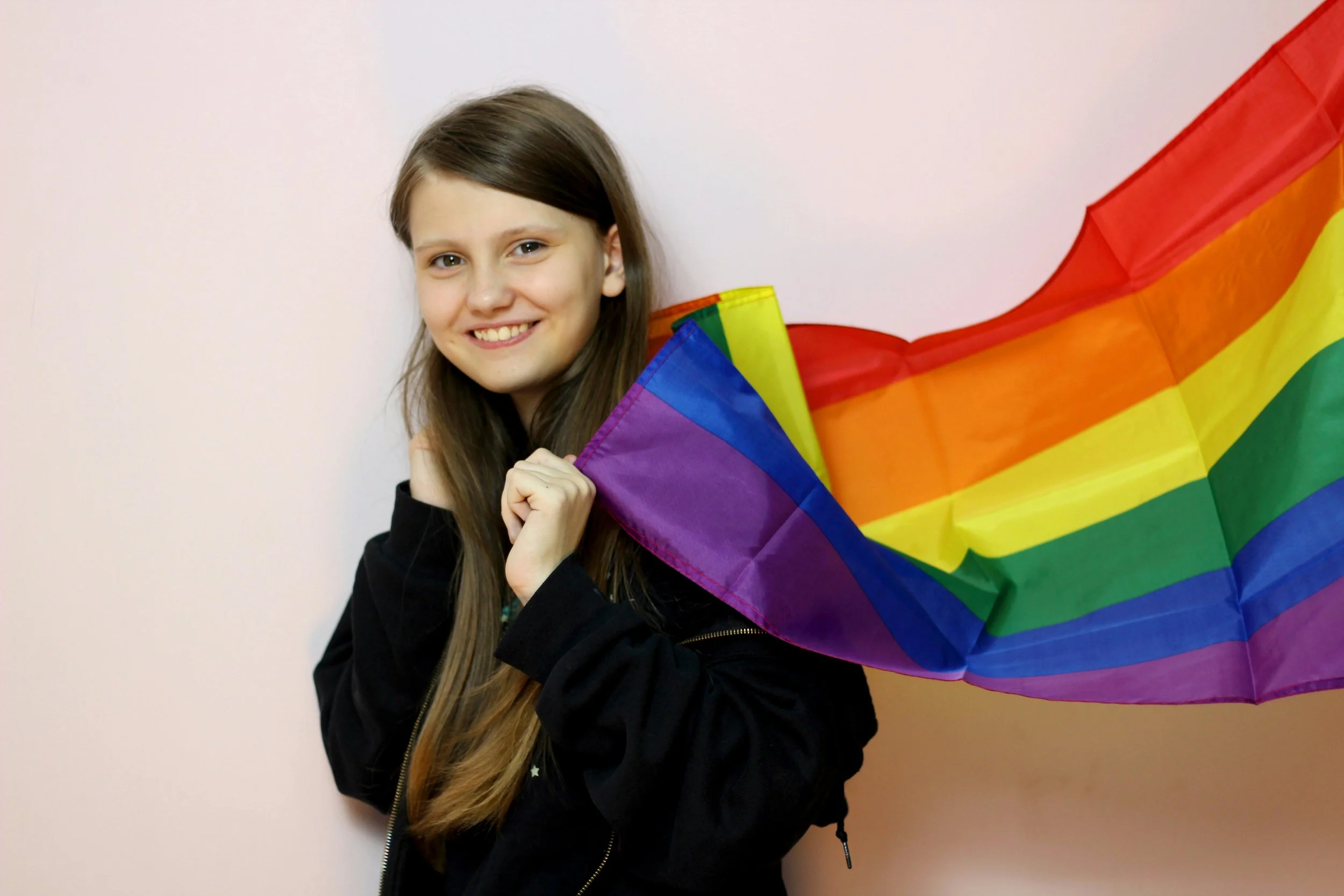 A smiling young girl holding a rainbow pride flag against a pink background.
