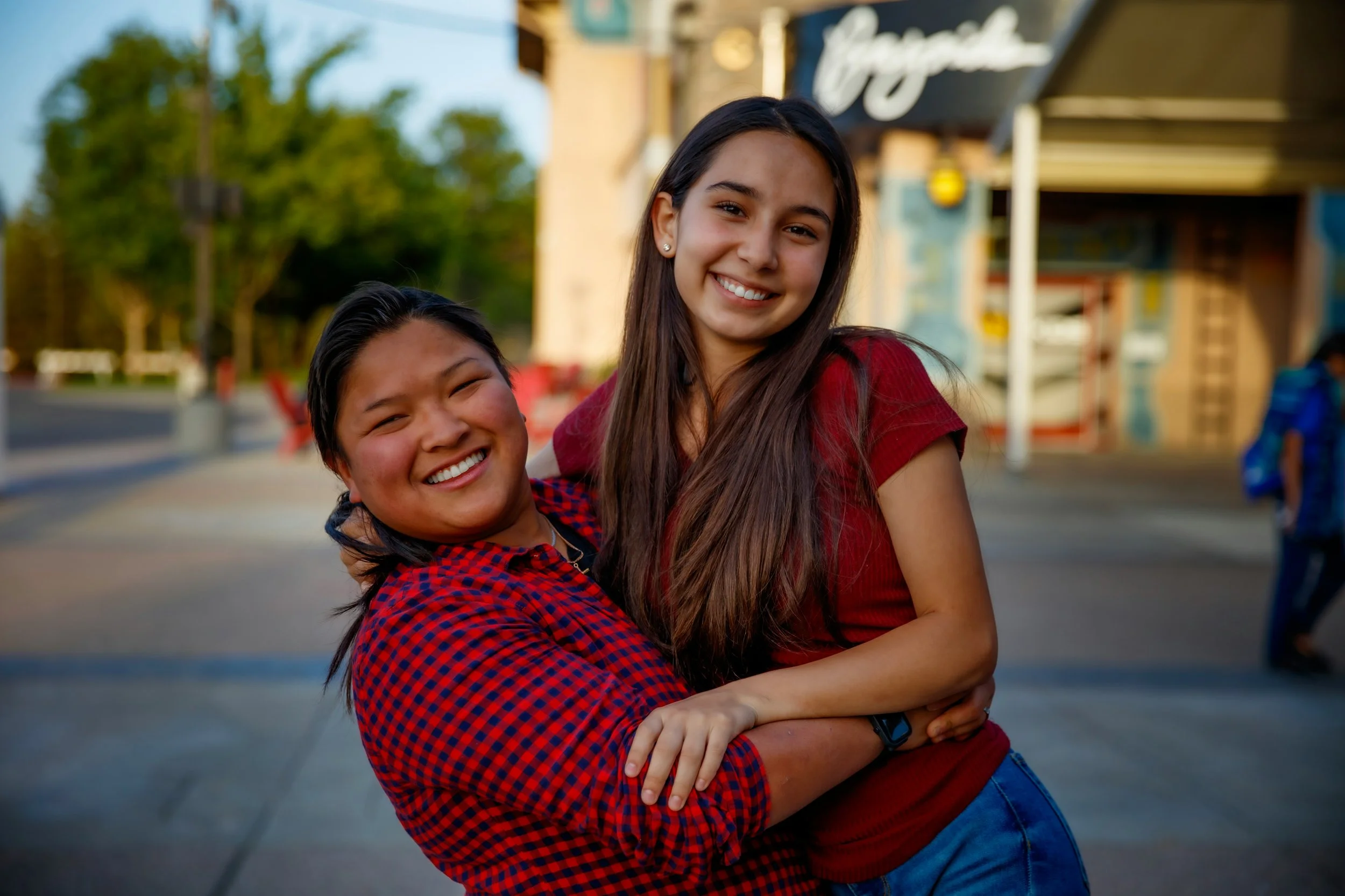 Two young women smiling and hugging outdoors in front of a building with trees in the background.