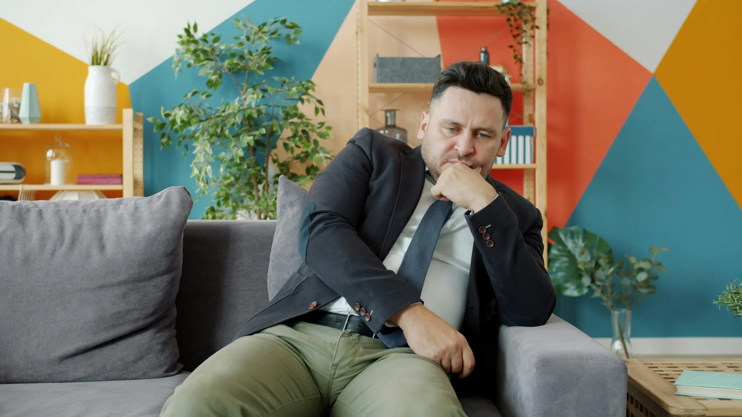 Man sitting on a gray couch, looking contemplative with one hand on his chin, in a colorful modern room with plants and shelves.