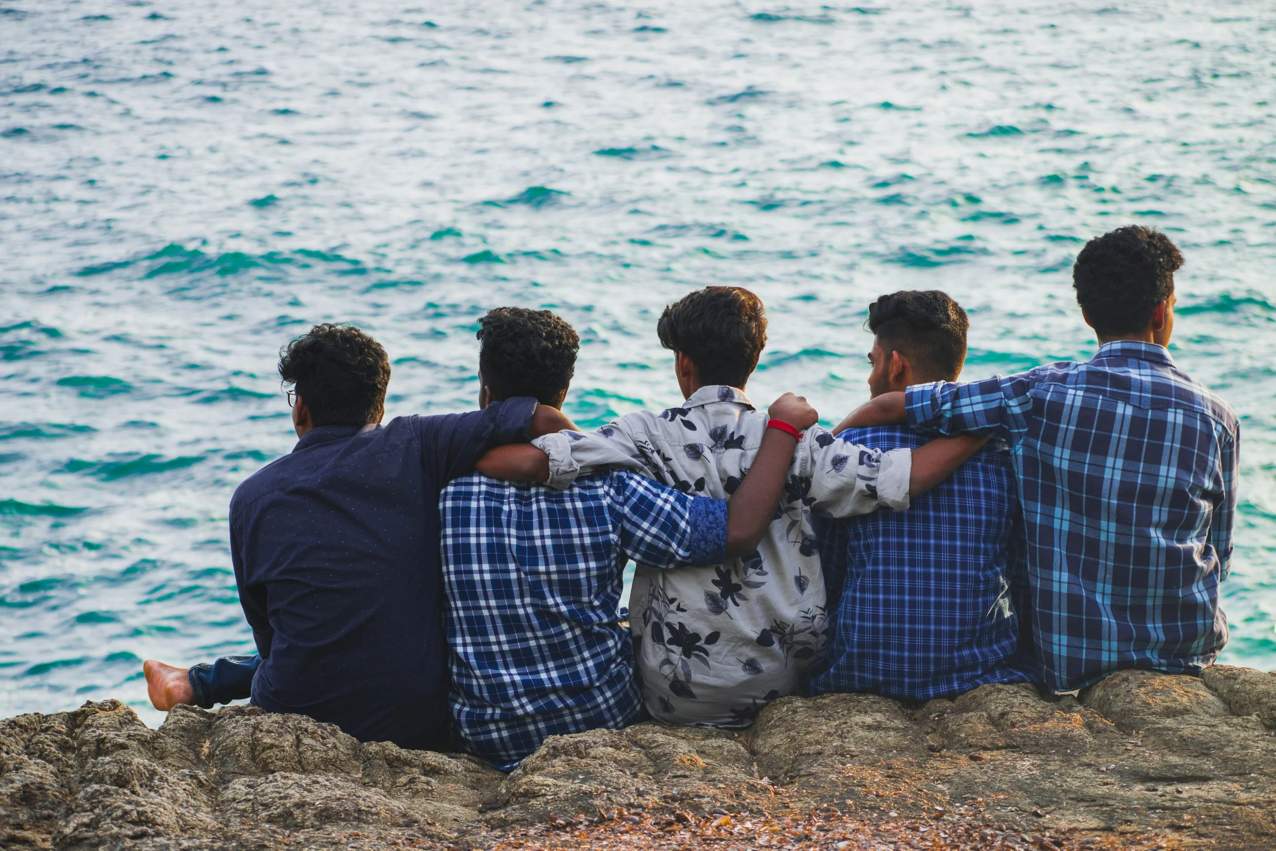 Five young men sitting on rocks by the water, watching the ocean, with their arms around each other's shoulders.