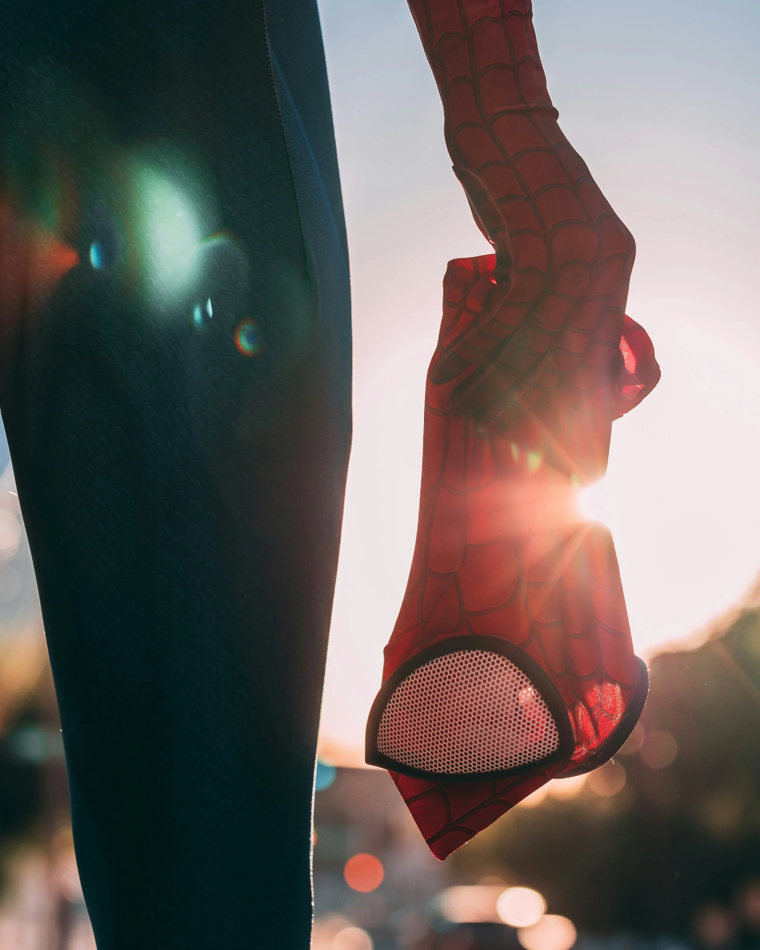 A person wearing blue athletic leggings holding a red Spider-Man themed mask with a built-in speaker, with a bright sun and lens flare in the background.
