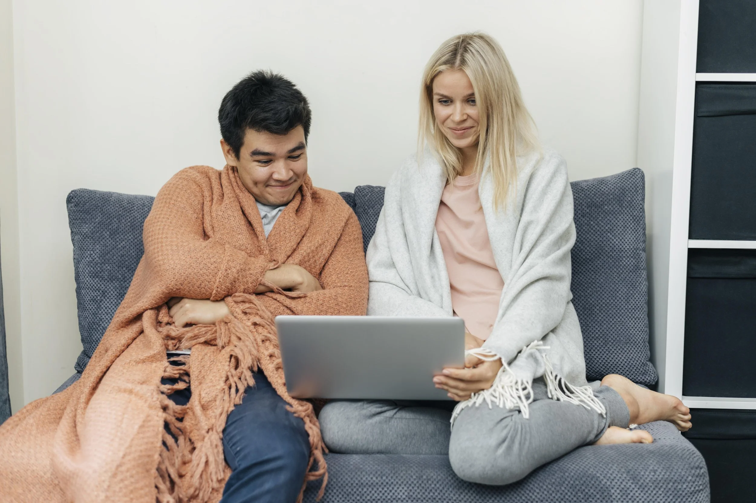 A young man and a woman sitting on a gray couch, looking at a laptop screen with smiles, wrapped in blankets in a cozy living room.