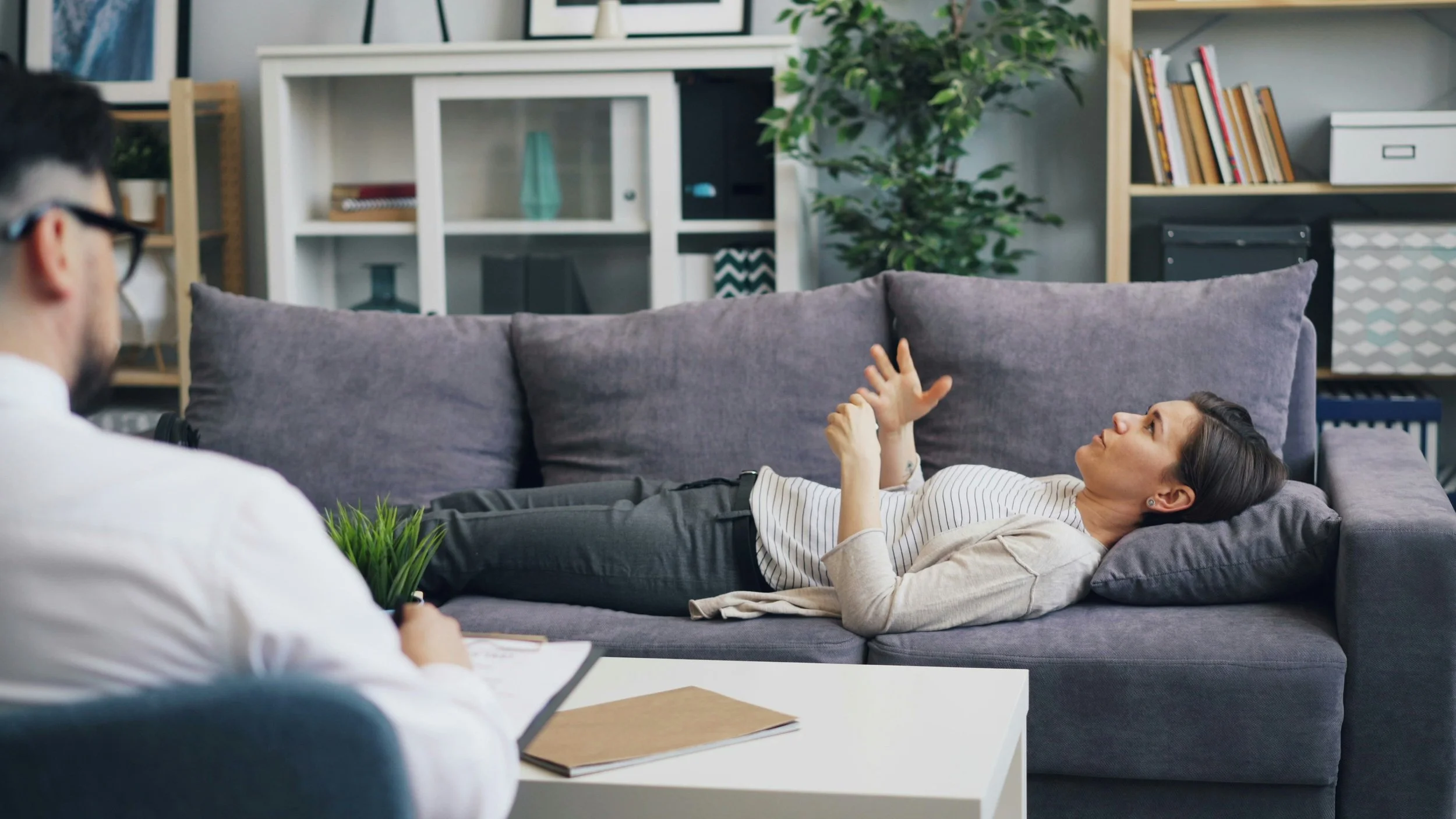 A woman lying on a sofa in a therapist's office, talking to a therapist who is taking notes.