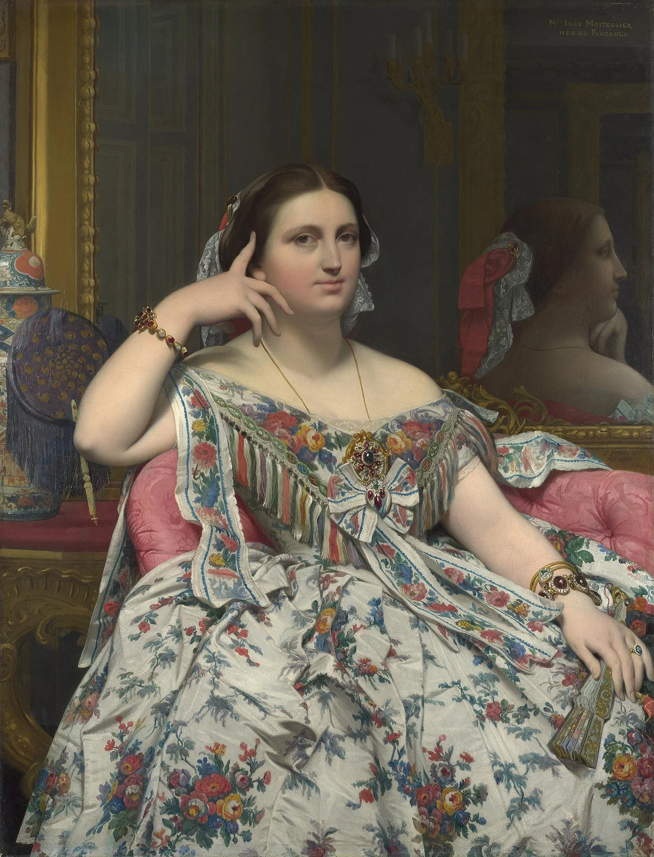 A portrait of a seated woman with dark hair, wearing a colorful floral dress and jewelry, with her hand resting on her face in front of a mirror reflecting her profile.