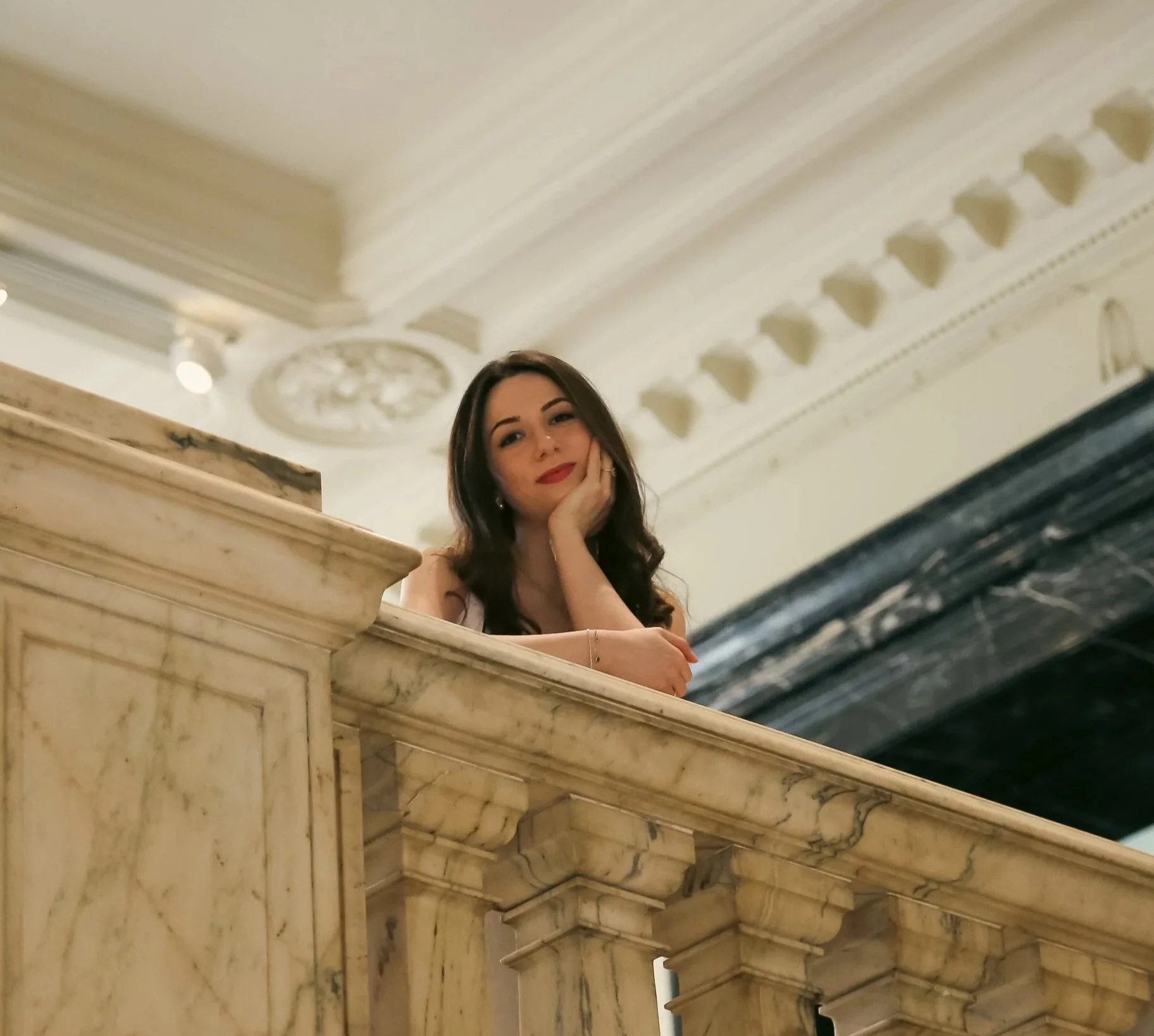 A woman with long dark hair resting her chin on her hand, leaning on a marble or stone bar, within an ornate interior with detailed crown molding and ceiling decorations.