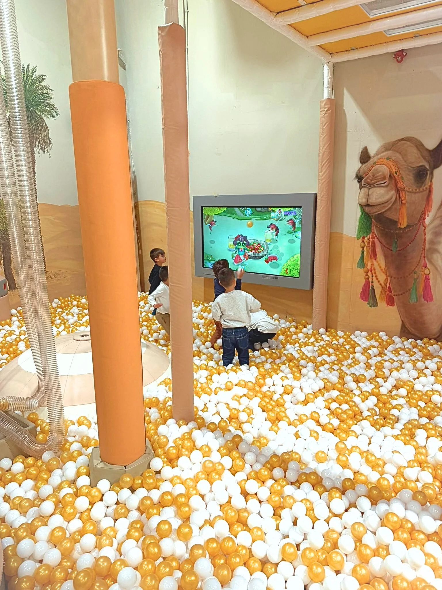 Children playing in a ball pit with white and gold balls, a large animated screen, and a giant camel statue with colorful decorations in an indoor play area.