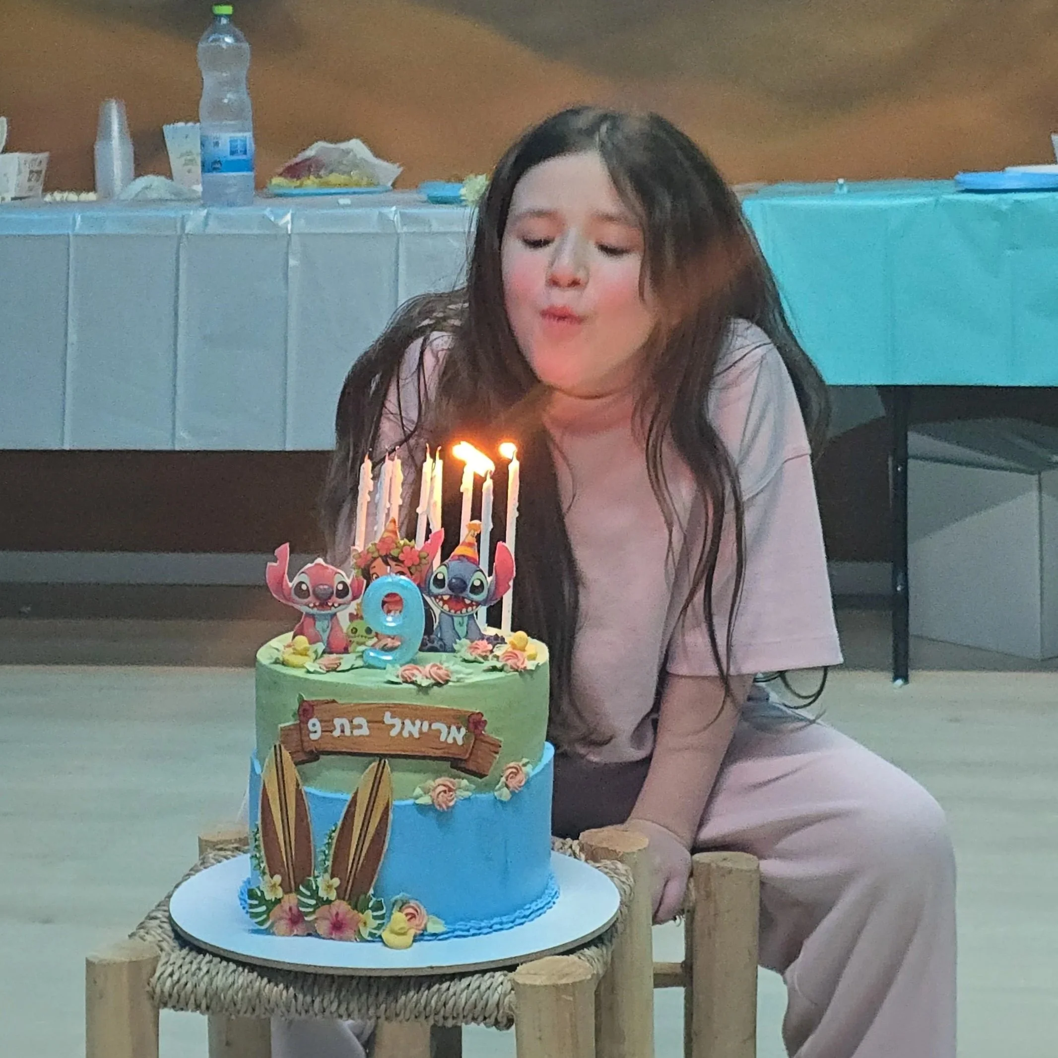 A young girl with long brown hair celebrates her birthday, blowing out candles on a colorful, themed birthday cake with cartoon character decorations, surrounded by birthday party items and an adult sitting nearby.