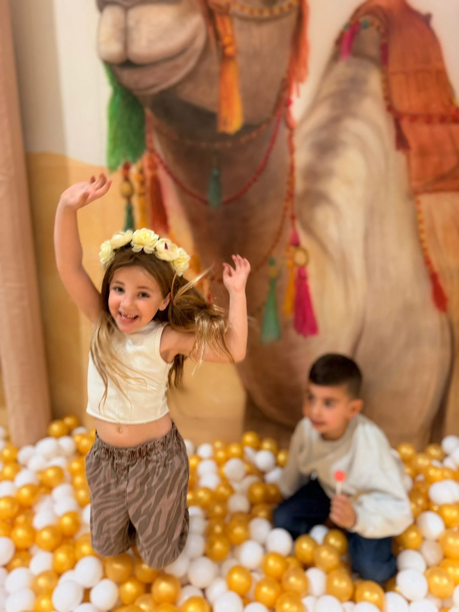 Children playing in a ball pit with gold and white balls, with a large colorful backdrop featuring a figure wearing traditional attire.