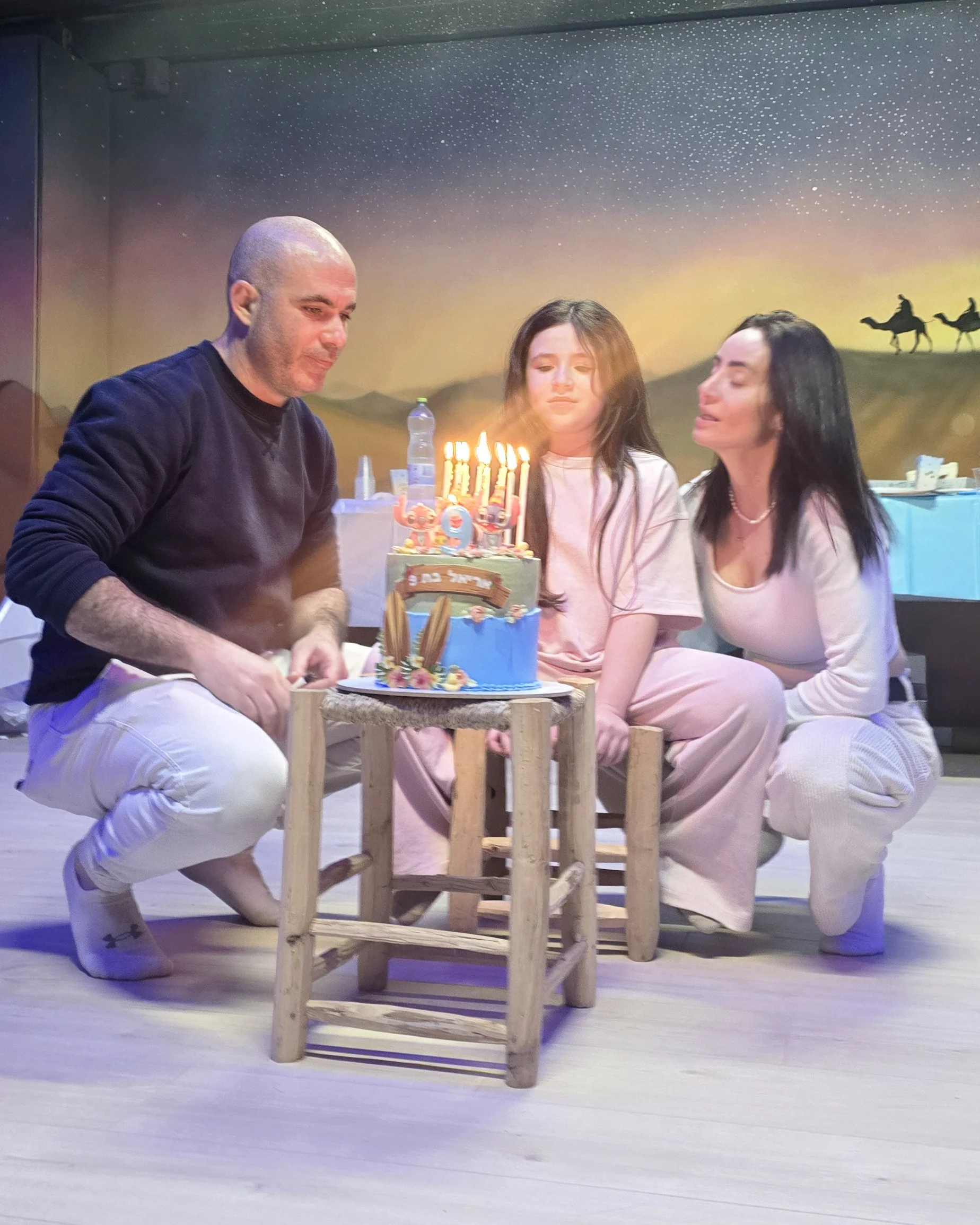 Three people, two women and one man, sit around a small table with a birthday cake with candles. The background features a painted mural of a desert scene with camels and a starry sky. The people appear to be celebrating a birthday.