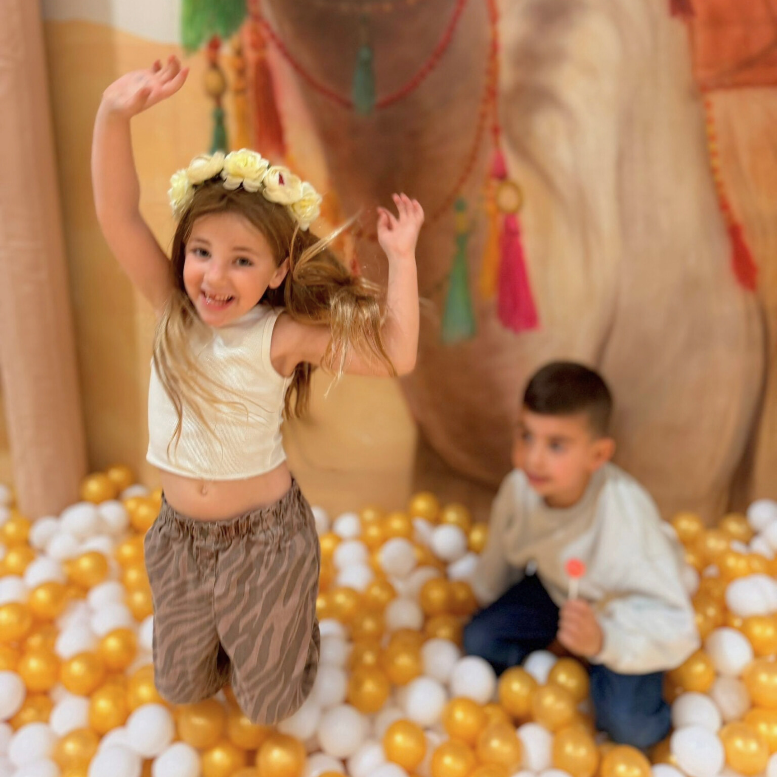 A young girl wearing a yellow flower crown playing in a ball pit with yellow and white balls, with another child partially visible in the background.