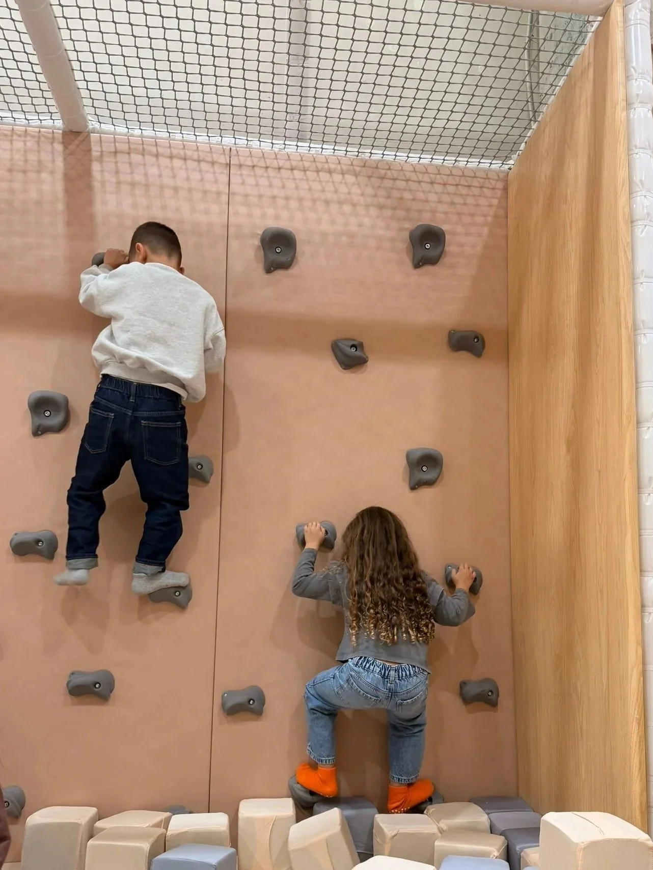 Two children climbing on an indoor rock wall with holds, one boy and one girl, in a play area with padded mat blocks on the ground.