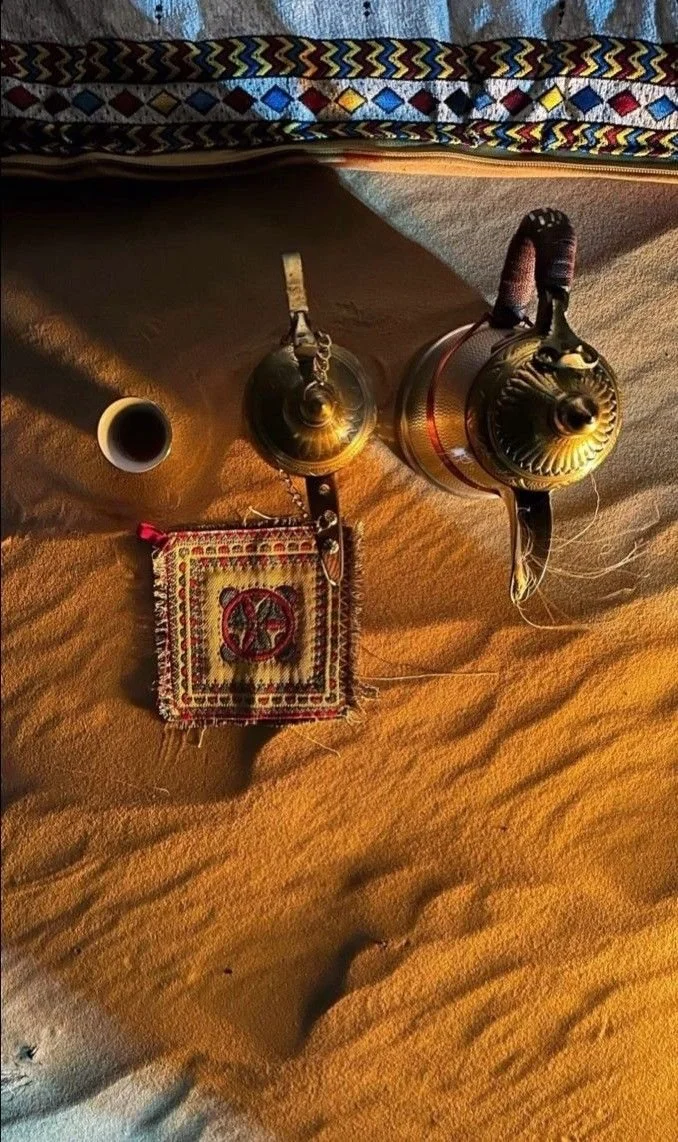 A desert scene with two traditional teapots, a cup of tea, a small decorative cloth, and a woven blanket at the top edge of the image.