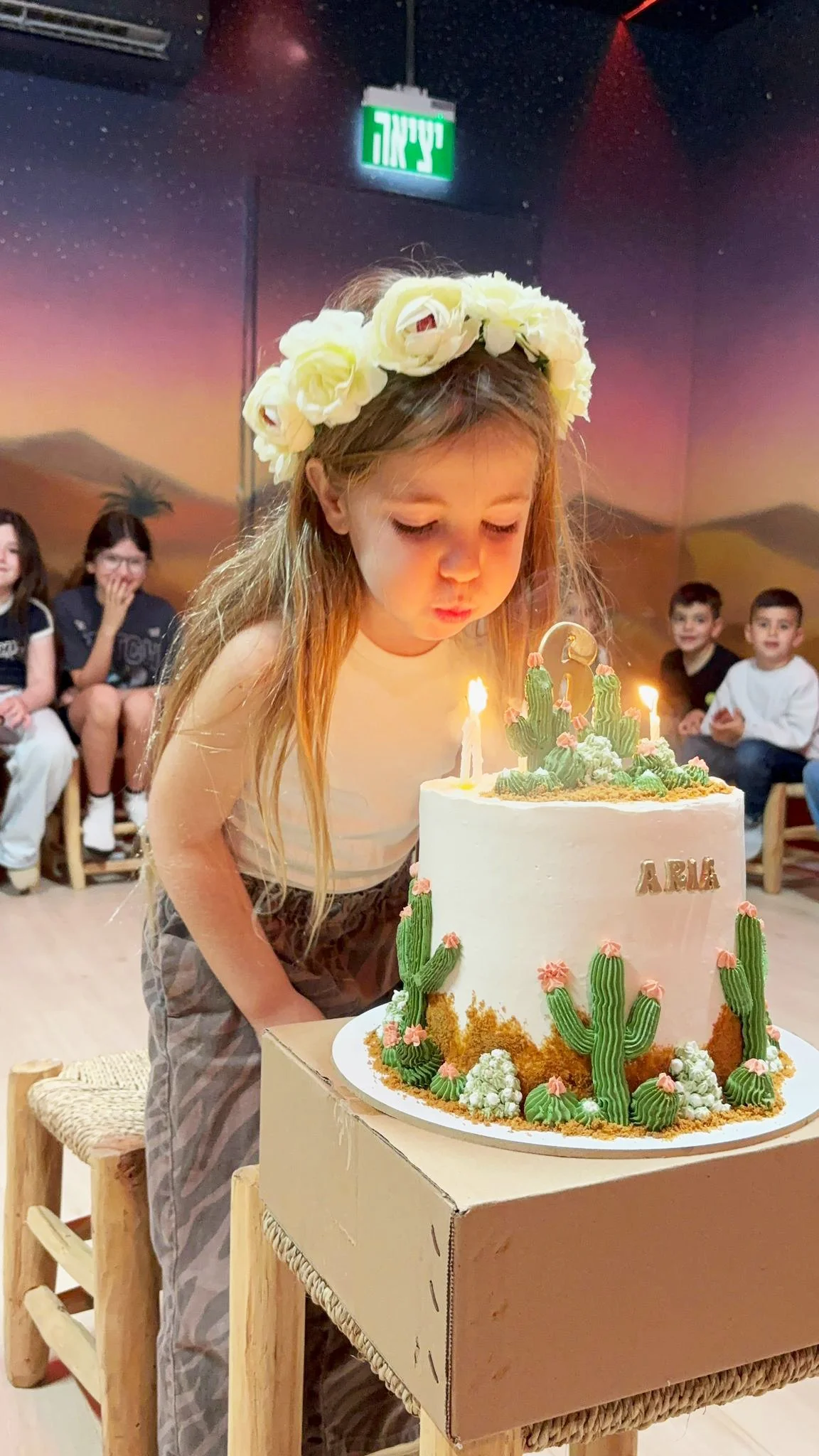 A young girl with a flower crown blowing out candles on her birthday cake decorated with cacti and desert plants, with people sitting in the background.
