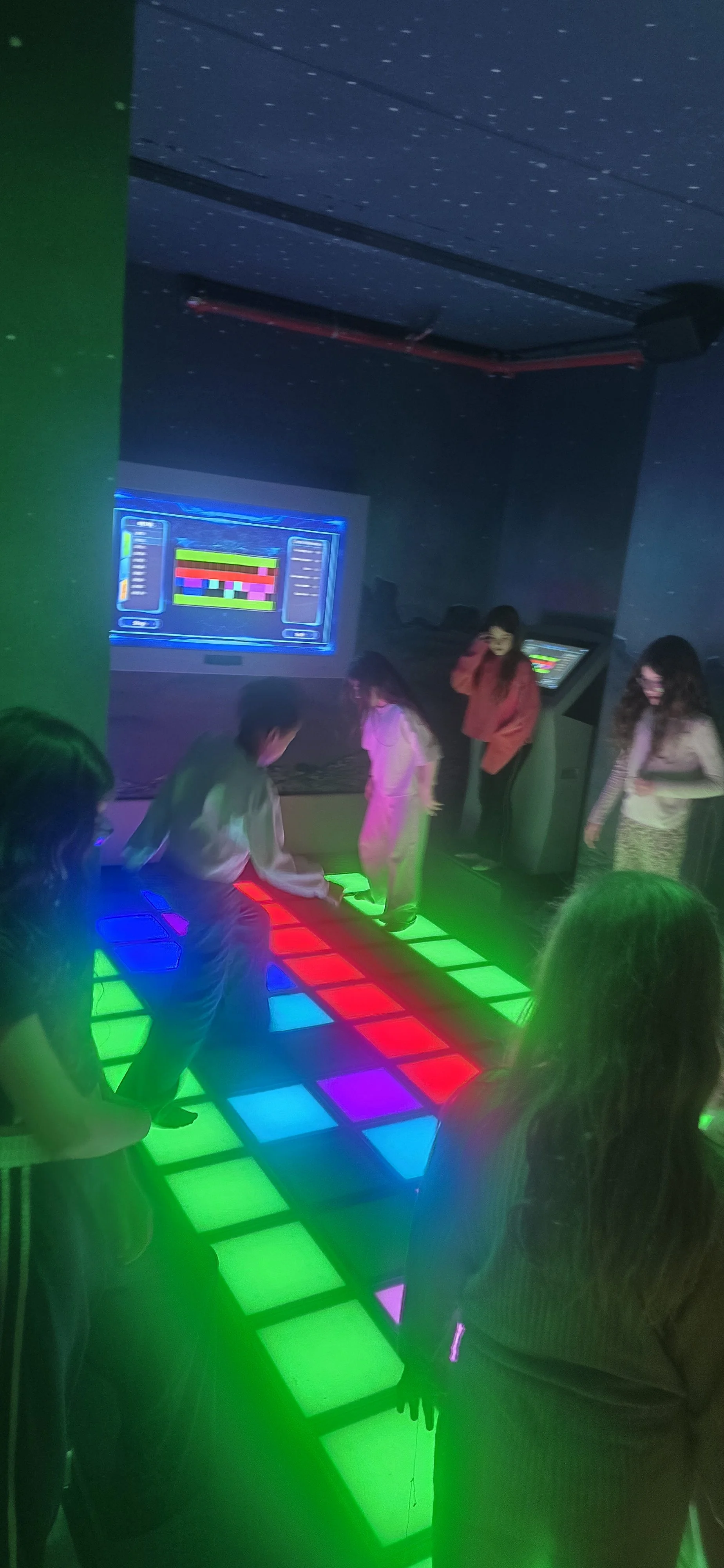People playing a colorful dance video game in a dark room with glowing screens and LED lights.