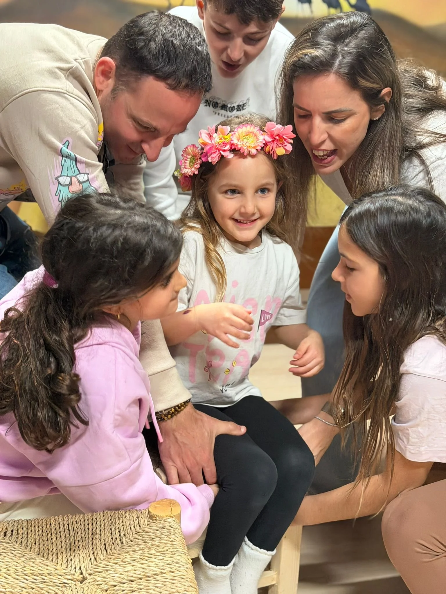 A group of people, including a young girl wearing a flower crown, are gathered closely together, engaging in a shared activity, possibly a discussion or craft, indoors.