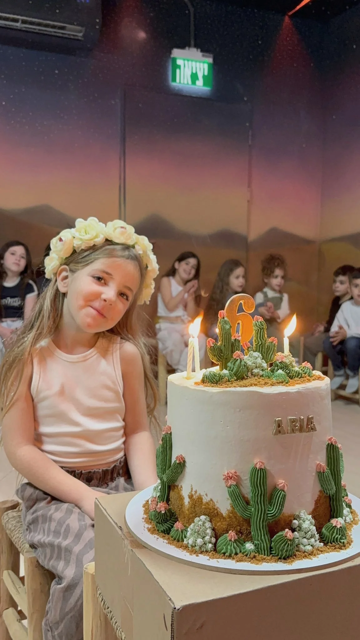 A young girl with a flower crown celebrates her birthday with a cactus-themed cake. She is sitting at a table in a room filled with children, and there are candles with the number 6 on the cake. The background shows a desert scene mural and other children seated nearby.
