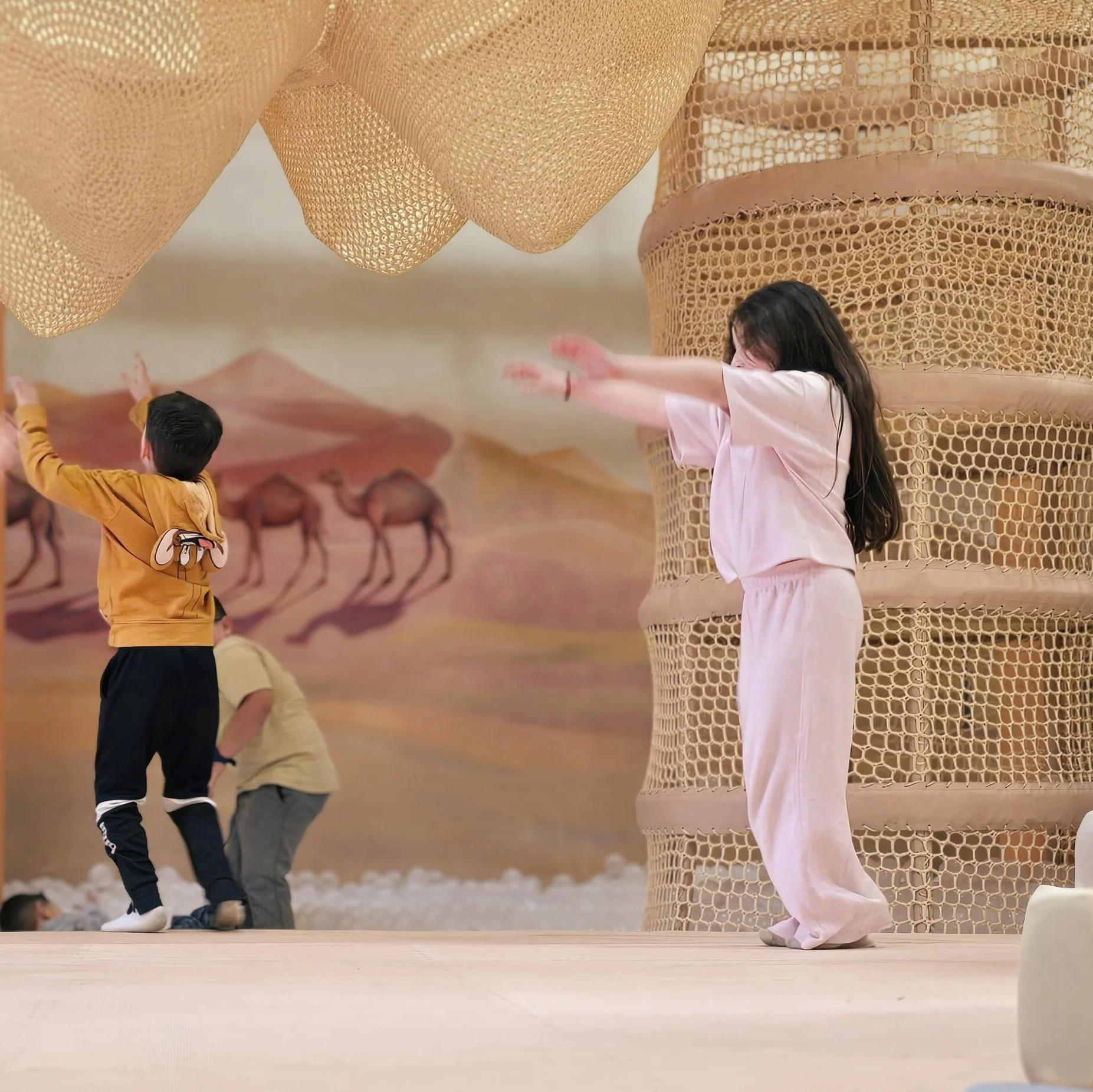 Children playing in an indoor playground with large woven structures resembling nests or cylinders, and a background mural of camels and desert landscape.