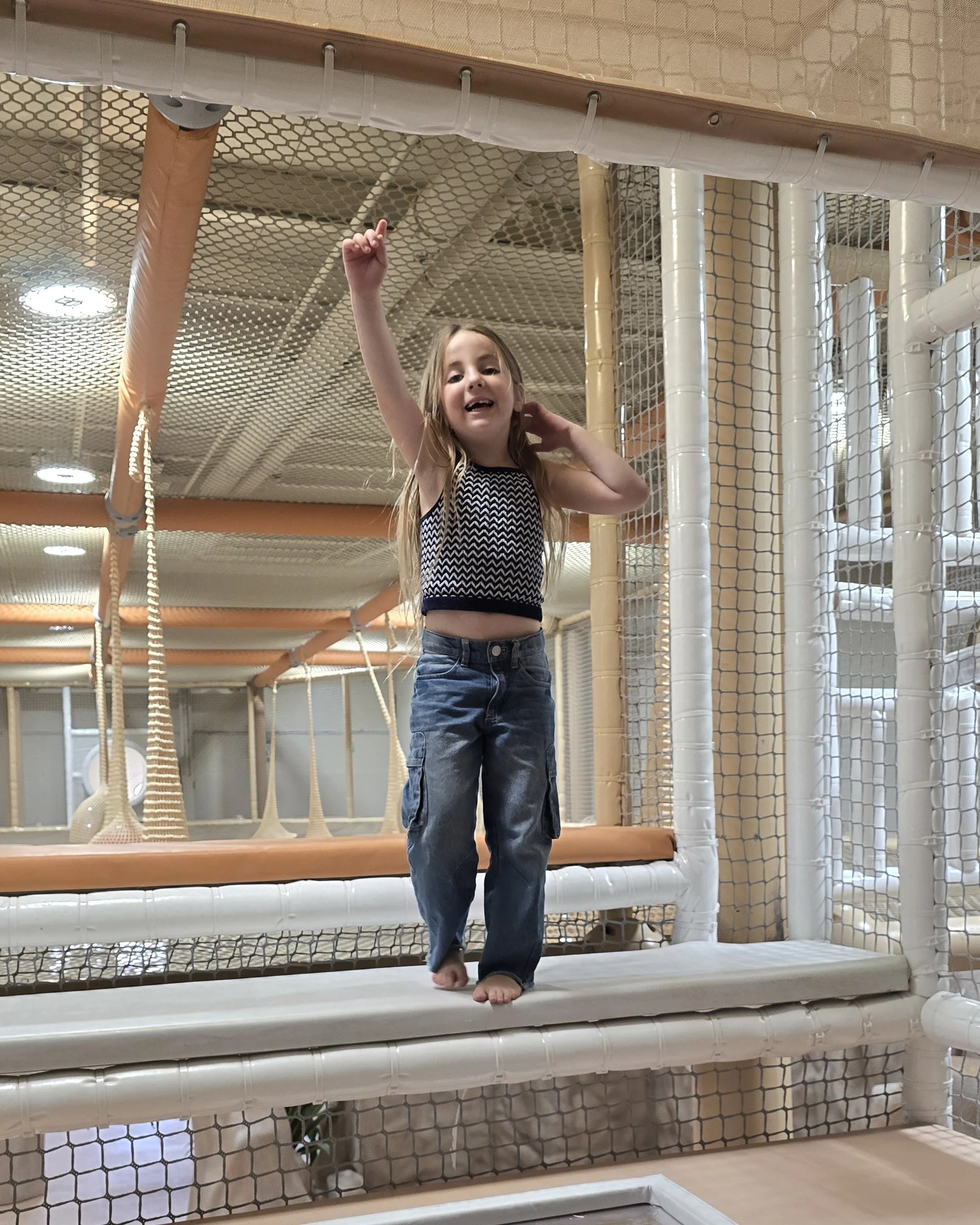 A young girl with long blonde hair wearing a black and white patterned top and blue jeans, standing barefoot on a padded surface in an indoor play area with climbing nets and wooden poles.