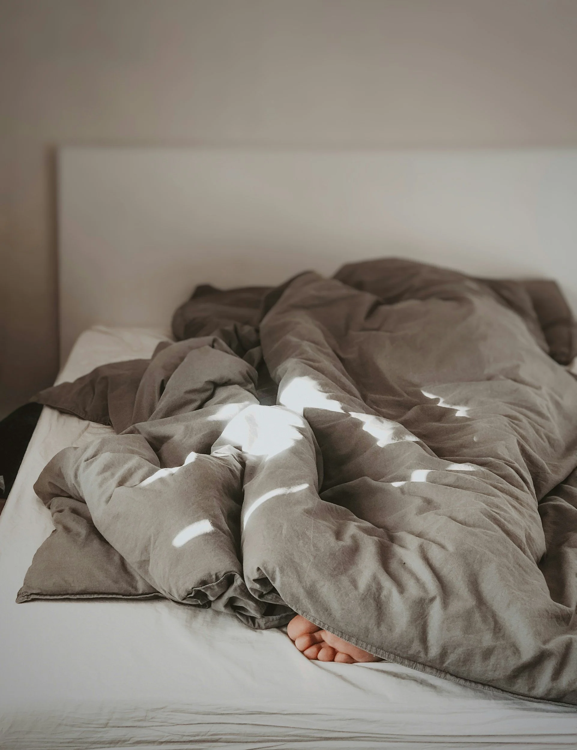Disheveled gray bedsheets and blankets on a bed with a white headboard, beige wall, and exposed foot peeking out from under the blankets.
