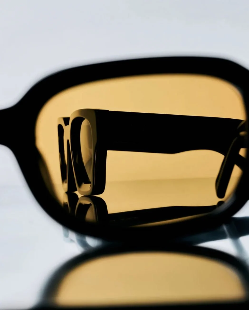 Close-up of a pair of sunglasses with yellow tinted lenses, reflecting the shape of a chair and part of a white wall.