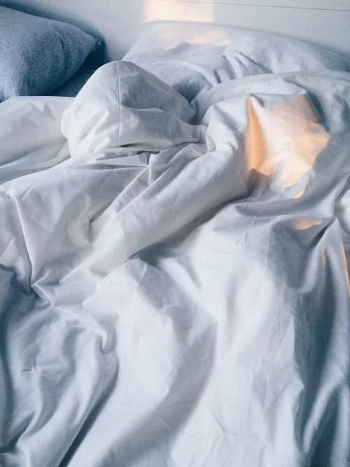 Disheveled white bed with crumpled sheets and pillows, illuminated by soft lighting.