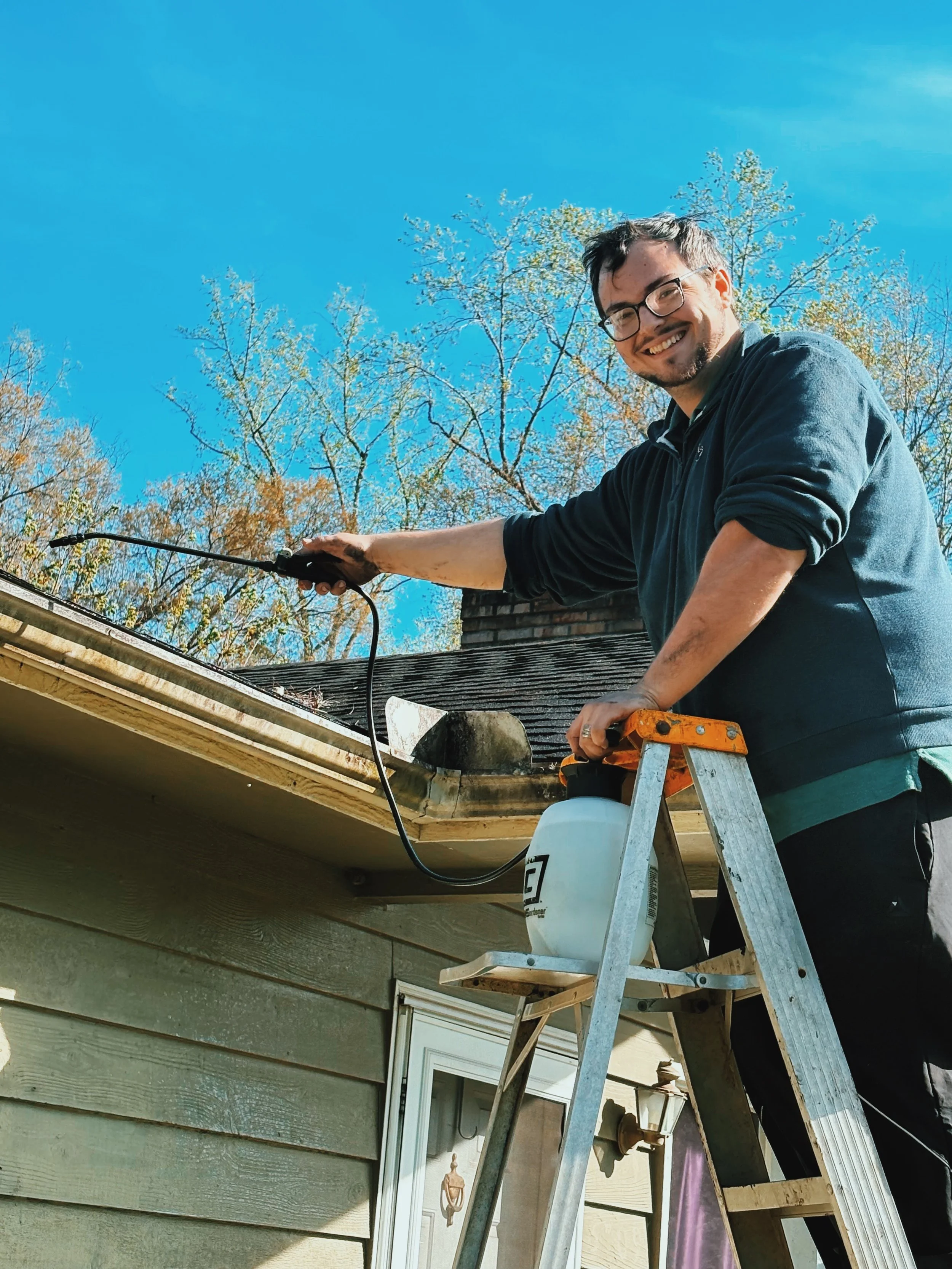 A man with glasses is standing on a ladder, using a spray nozzle connected to a sprayer to clean or treat the roof of a house. He is smiling, outdoors, with trees and a blue sky in the background.