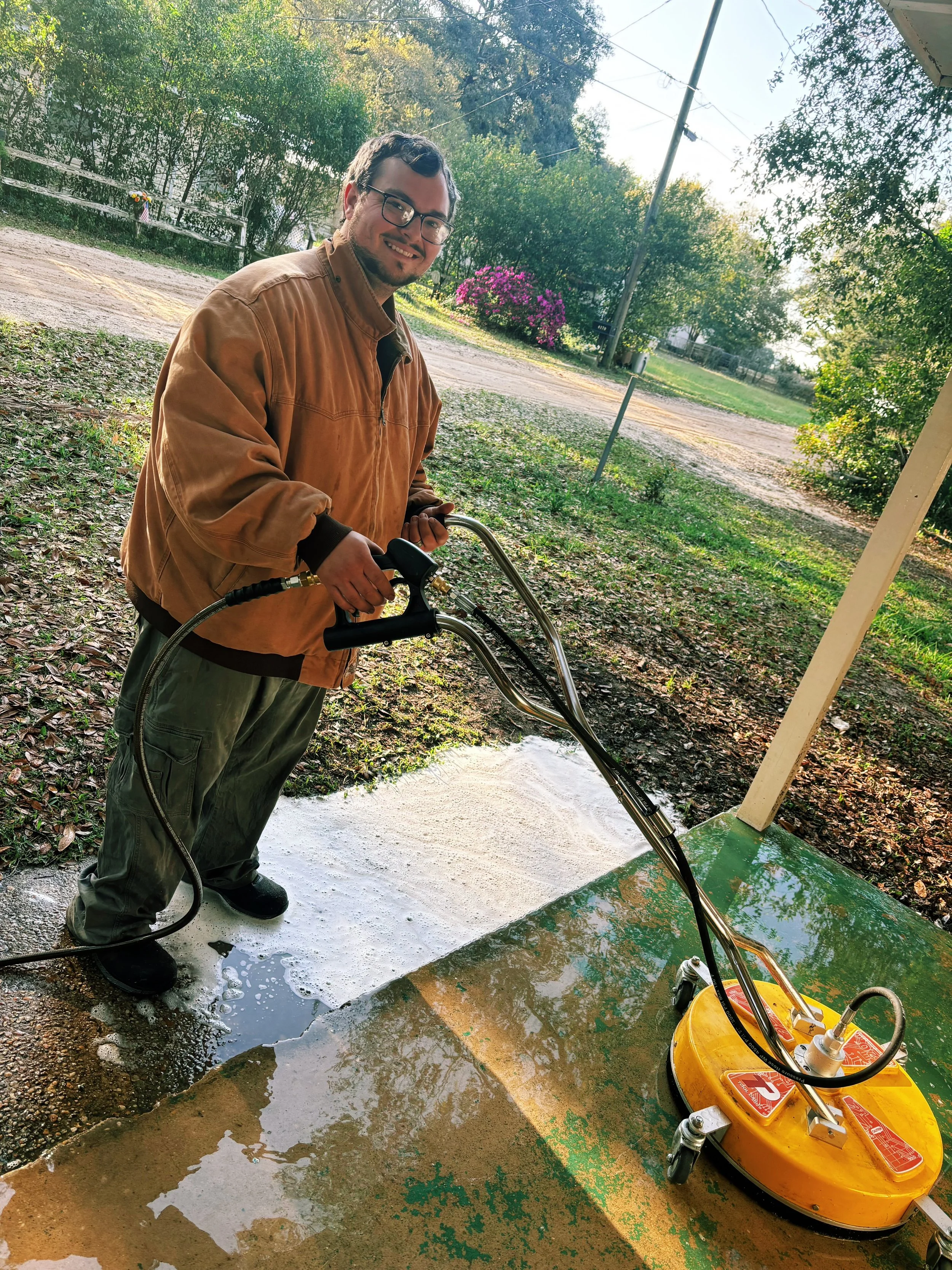 A man is washing a concrete surface outdoors using a pressure washer, standing on wet concrete with soap suds, surrounded by trees and greenery.