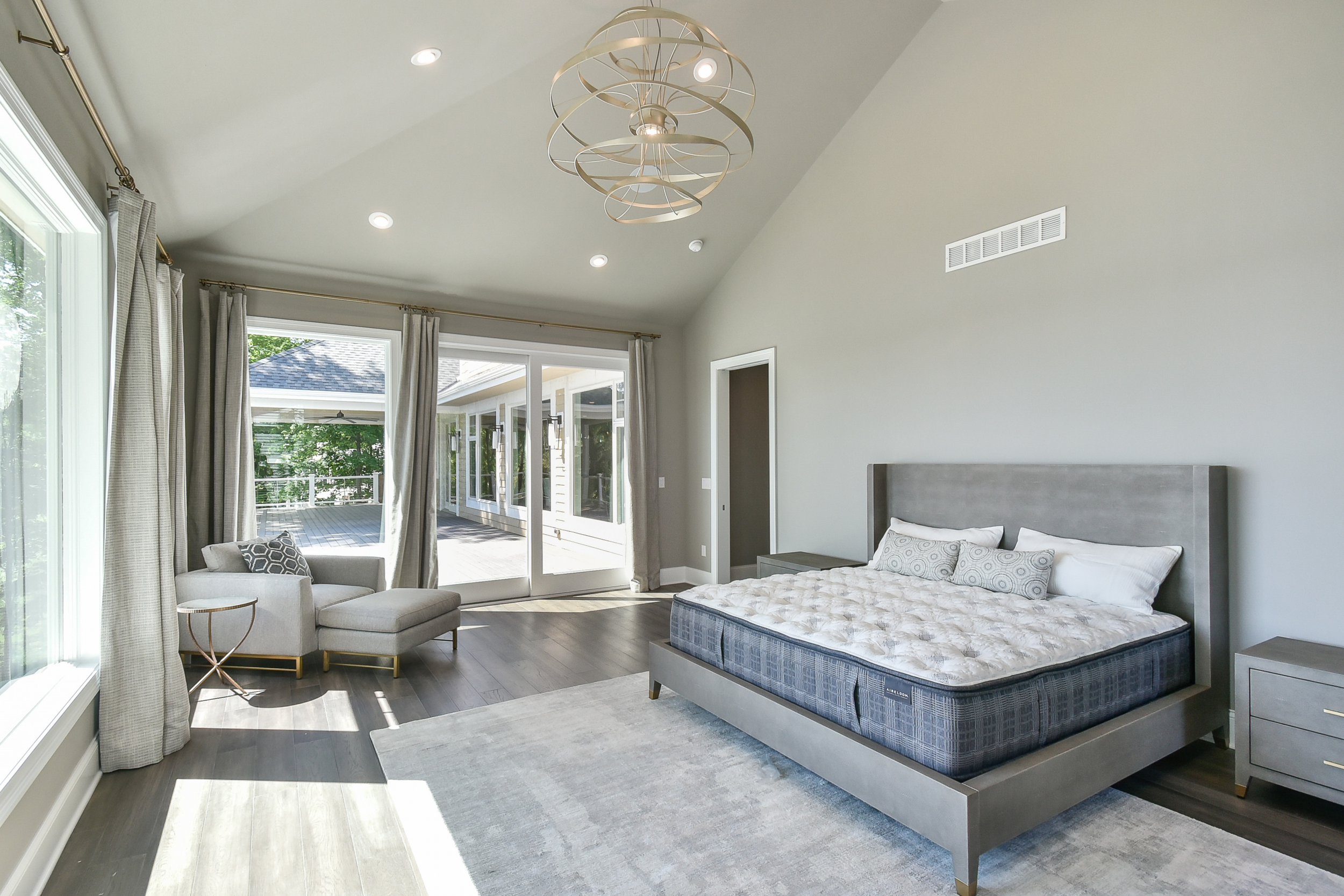 A modern bedroom featuring a large bed with a gray headboard, a mattress, and decorative pillows. There are two gray nightstands on each side, a beige armchair with an ottoman and a geometric pillow, and large glass sliding doors that lead to an outd