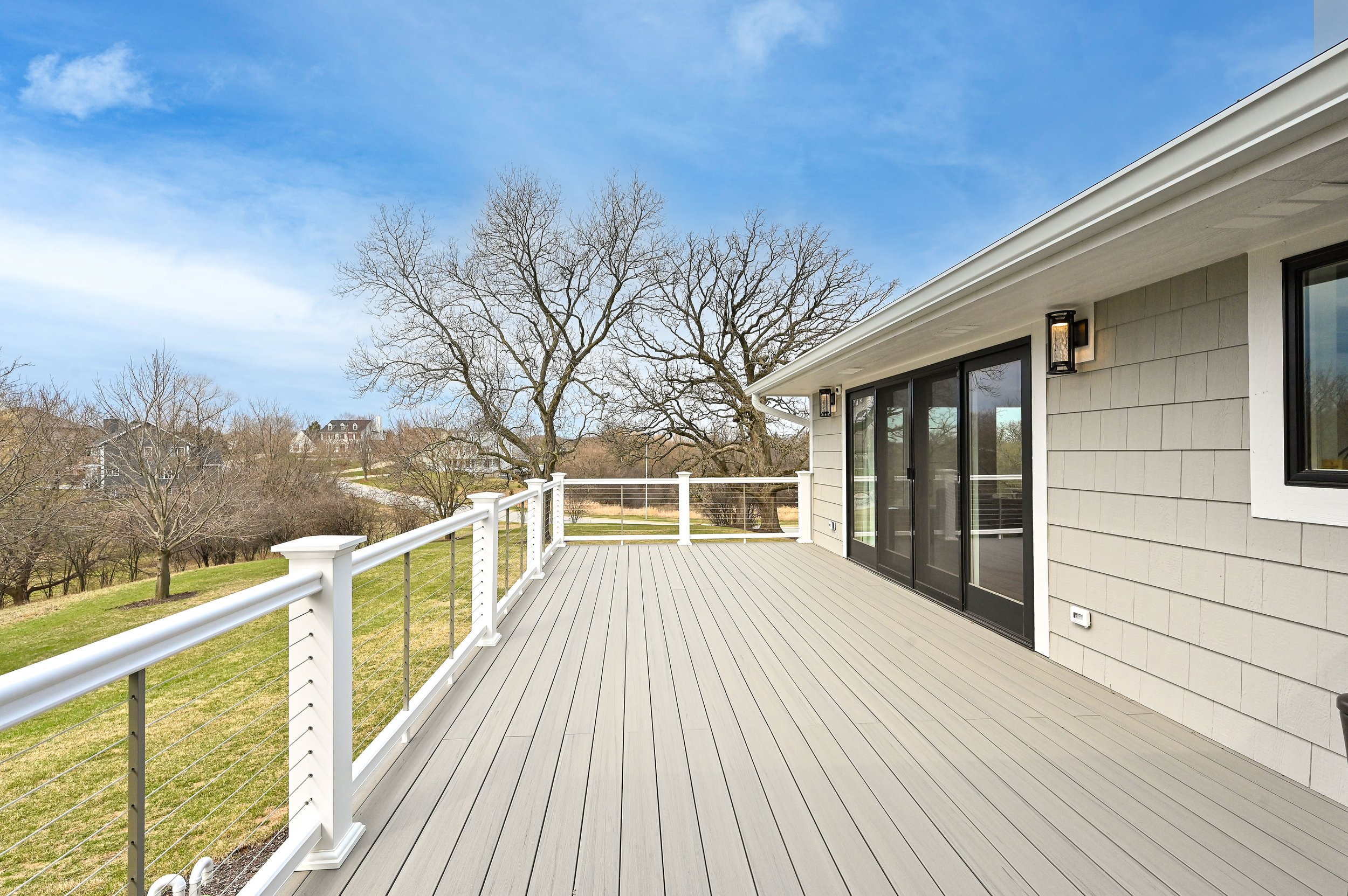 A spacious outdoor deck attached to a house with sliding glass doors, outdoor lighting, and a white railing, overlooking a landscape with leafless trees and a clear blue sky.