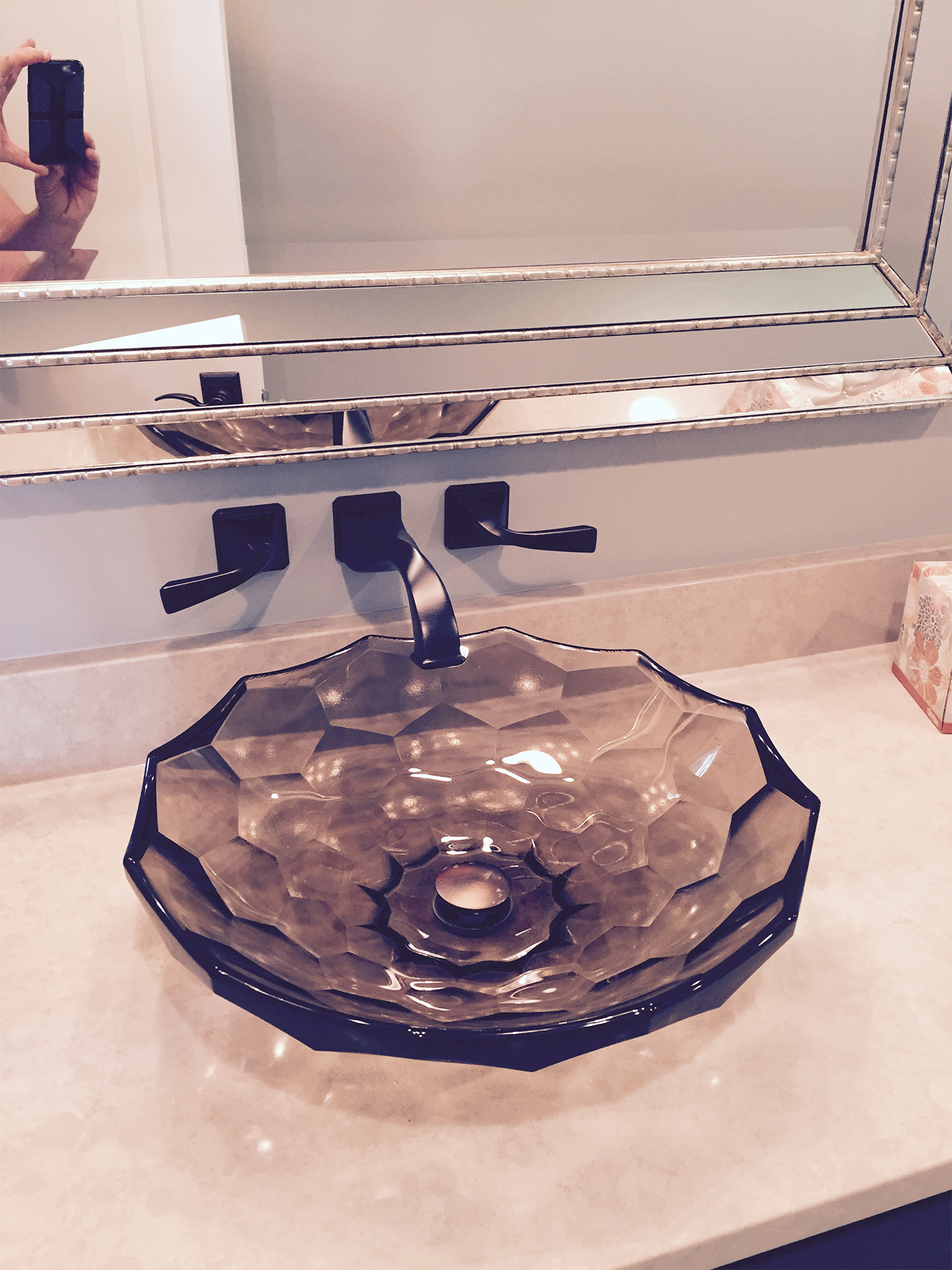 A decorative glass sink with a honeycomb pattern sits on a beige countertop in front of a large mirror, with a black faucet above it and black handles on either side.