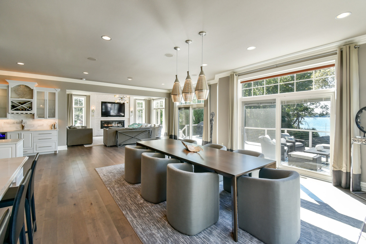 Open-concept living and dining area with large windows and sliding glass door leading to a balcony with outdoor seating, view of water and trees, modern furnishings, pendant lights over dining table, fireplace with mounted TV, and kitchen in backgrou