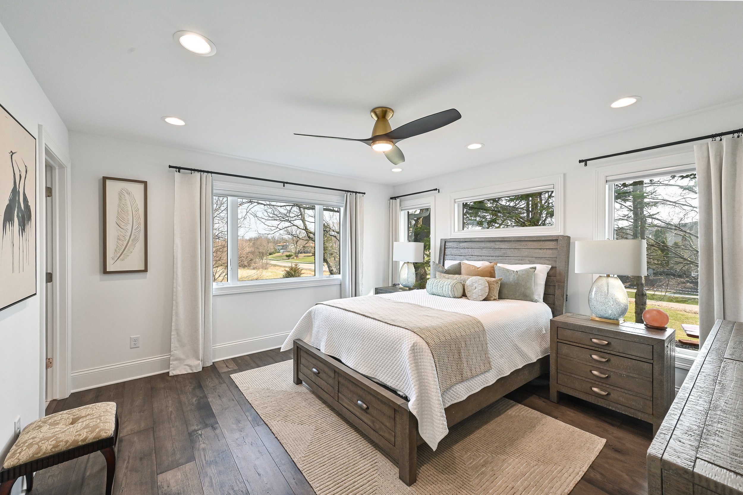 A modern bedroom with large windows, a gray wooden bed with white bedding, matching nightstands with lamps, and framed art on the white walls.