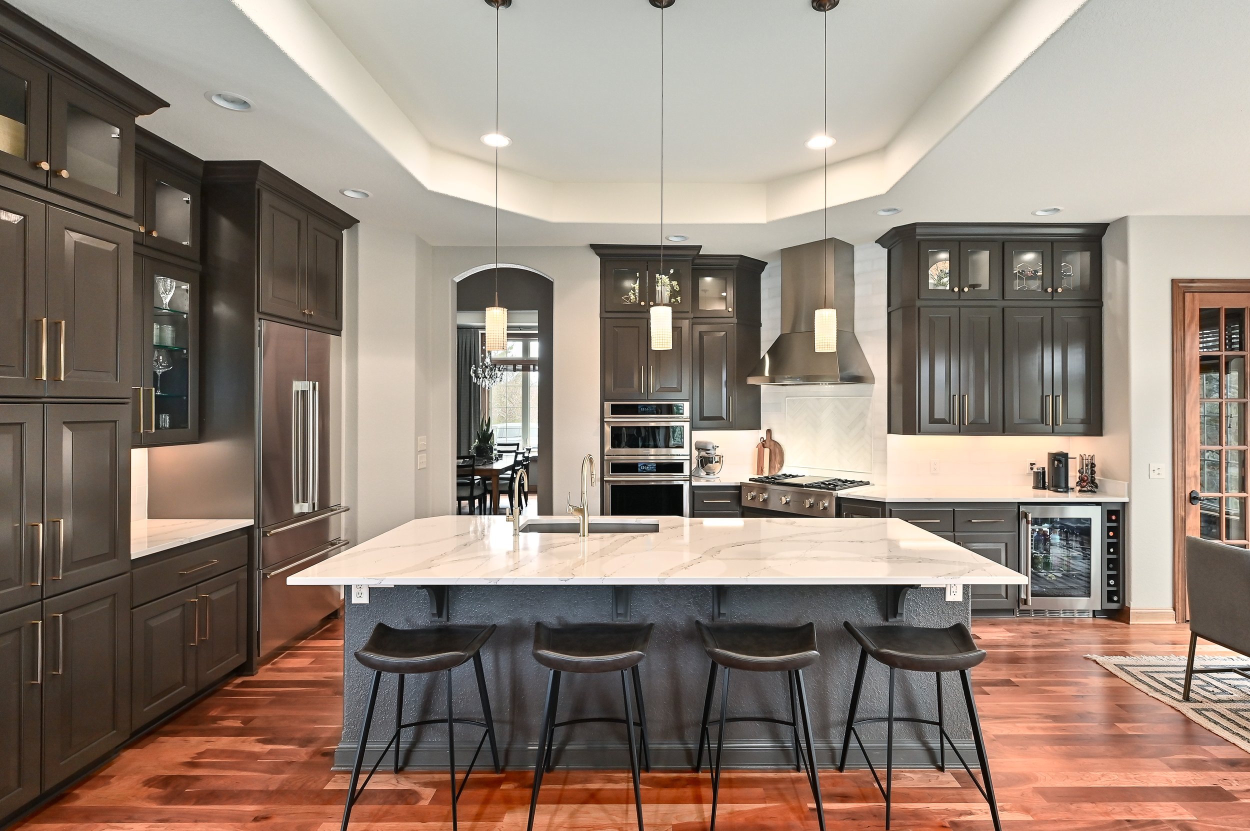 Modern kitchen with dark gray cabinets, white marble island, and hardwood floors. Includes stainless steel refrigerator, oven, and range hood, with pendant lights hanging above the island.