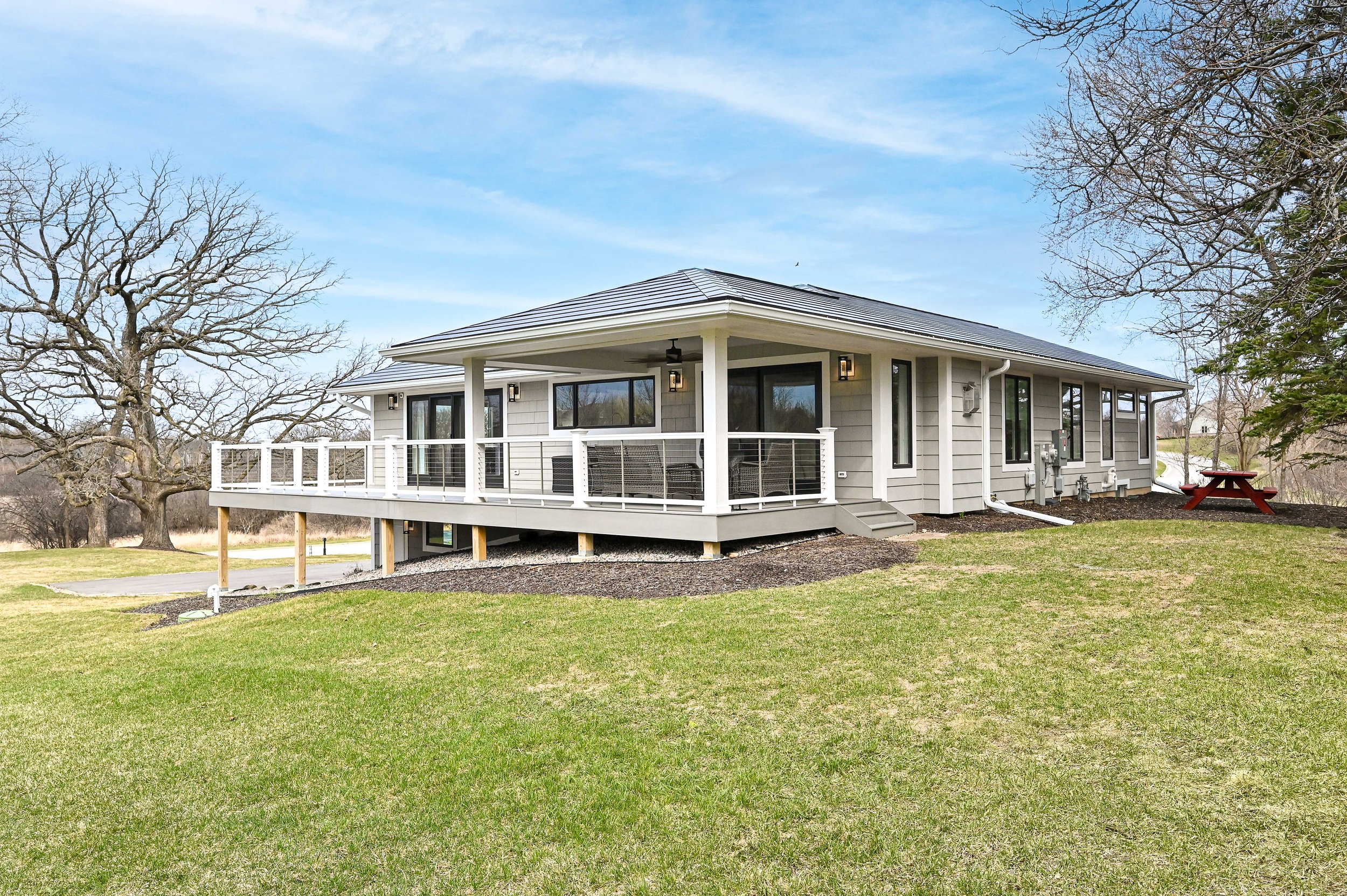 Modern house with a large wooden deck, white railing, and multiple outdoor light fixtures. Surrounded by a grassy lawn and trees, with a clear blue sky overhead.