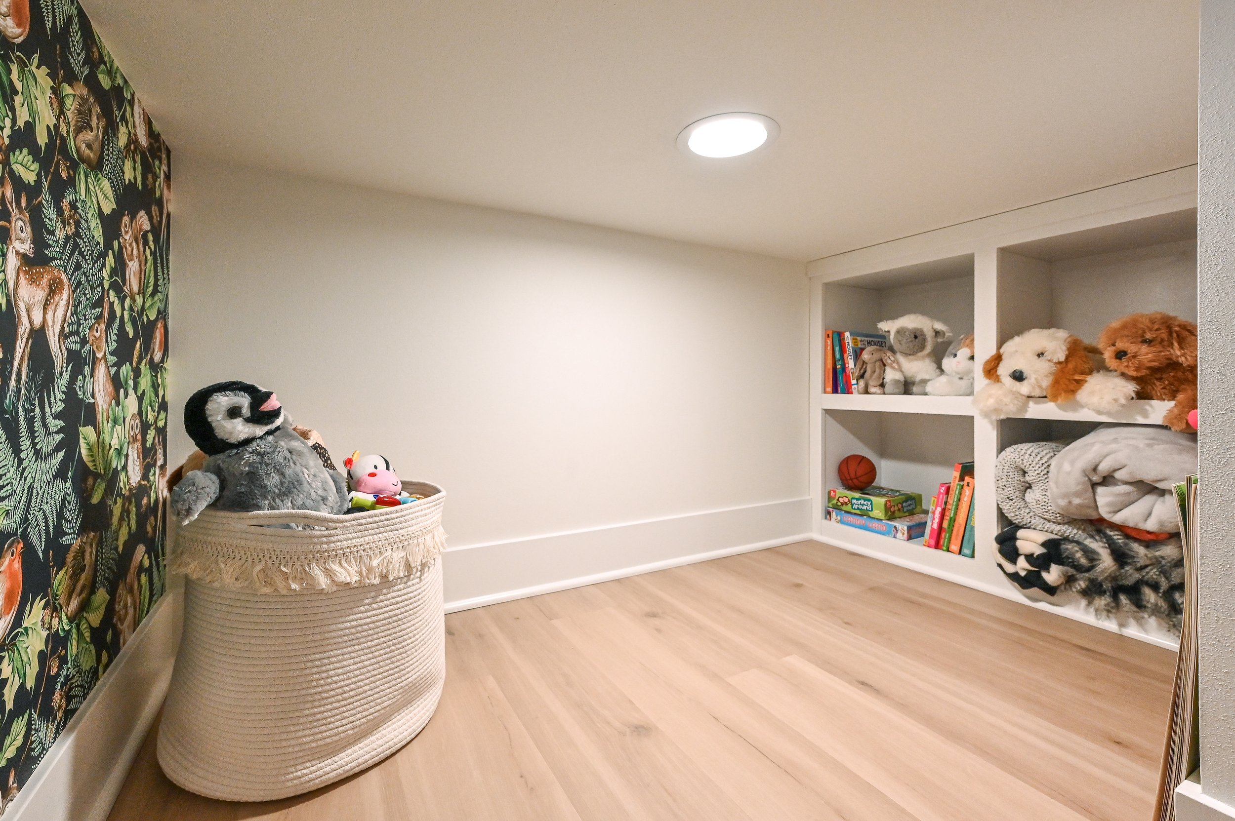 A small, brightly lit toy storage room with plush toys on shelves, a laundry basket with stuffed animals, colorful books, and a decorative wall with jungle animal print.
