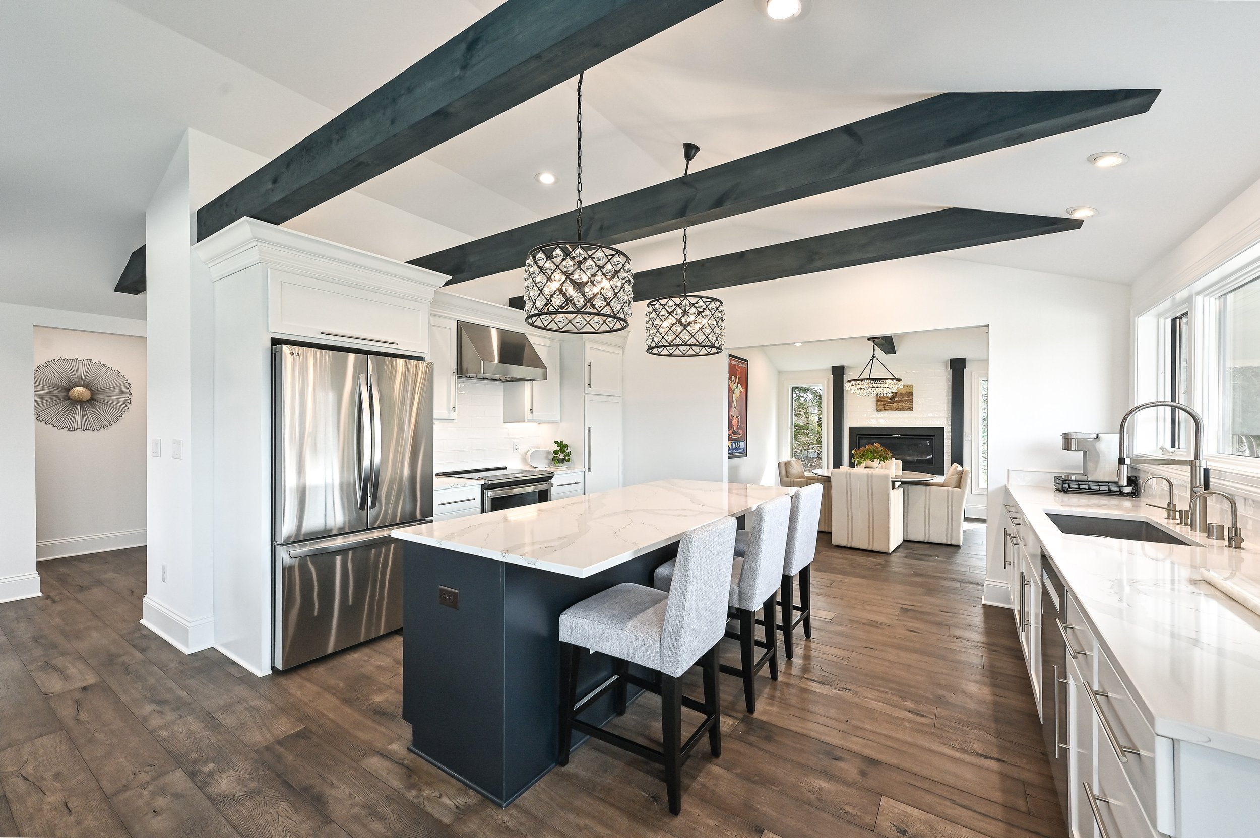 Modern kitchen with white cabinets, a large marble island, stainless steel refrigerator, black beams on the ceiling, and a view into a living room with a fireplace and white chairs.