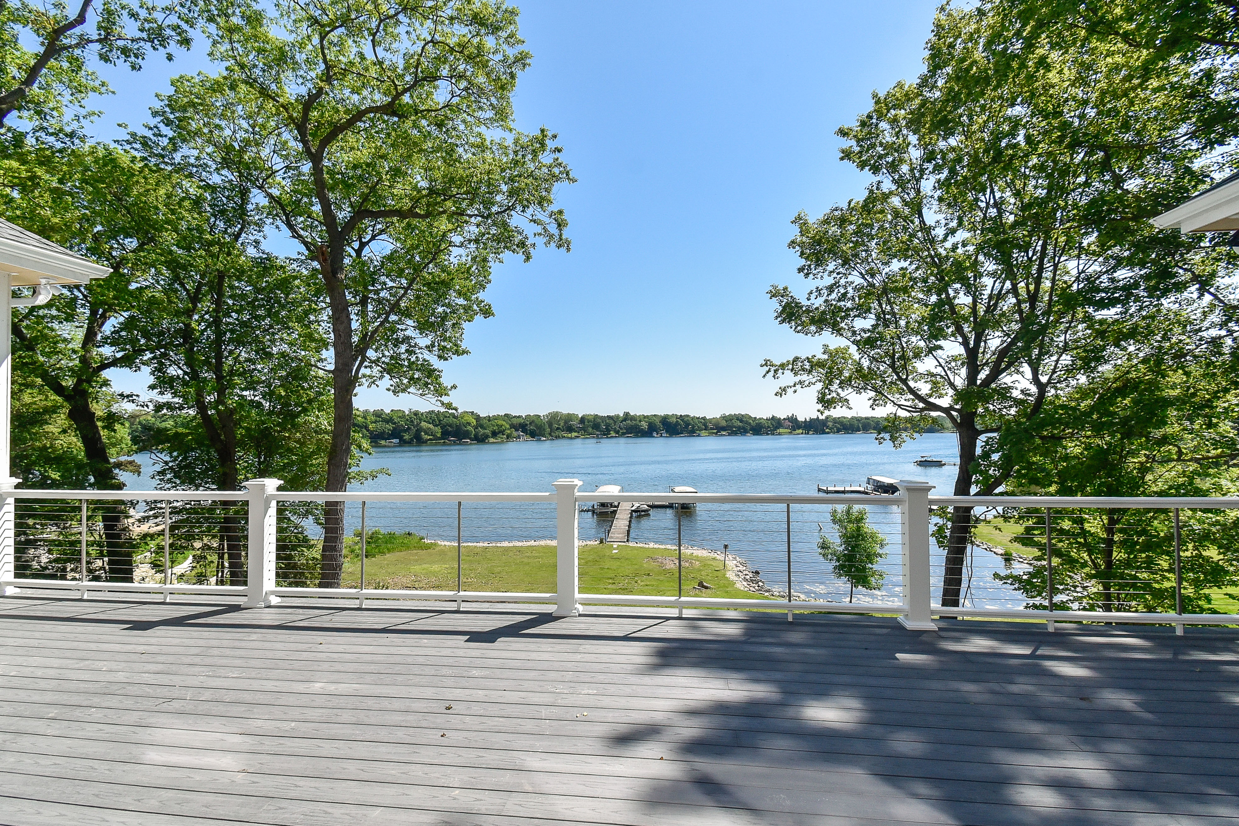 A view of a lake seen from a wooden deck with white railing, surrounded by green trees under a clear blue sky.