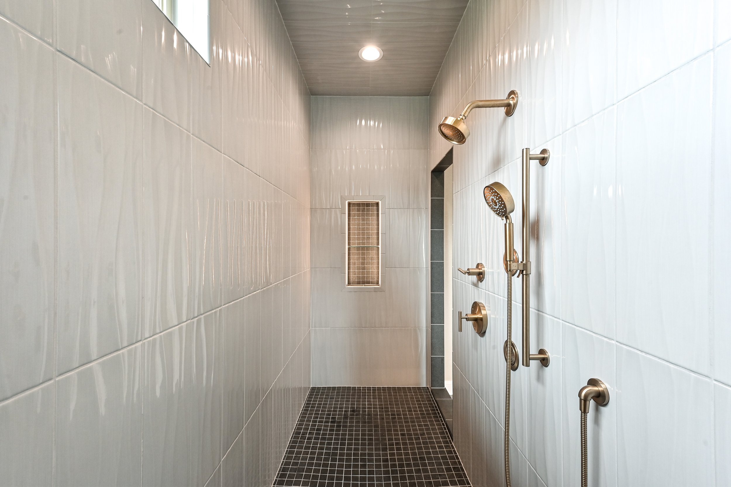 Empty walk-in shower with white tiled walls, black tiled floor, and multiple shower heads and controls mounted on the right wall.
