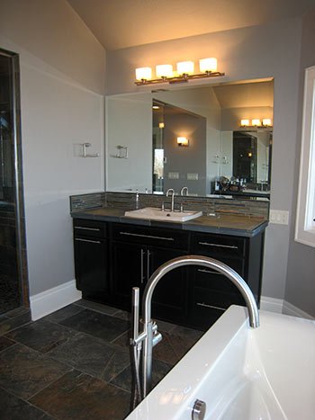 Bathroom with a large mirror, vanity with dark cabinets, and four light fixtures above. Part of a bathtub with a chrome faucet is visible in the foreground.