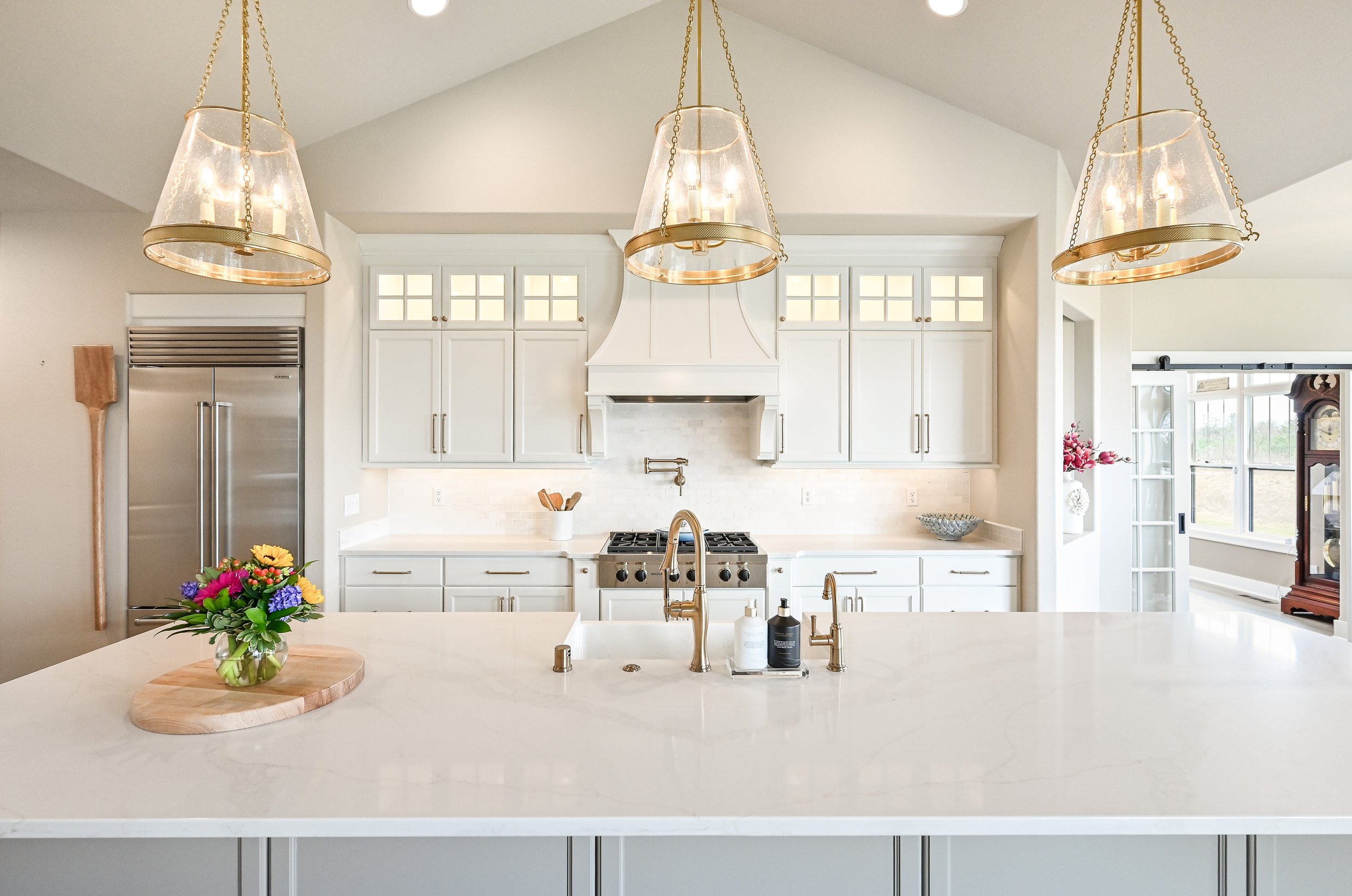 Bright, modern kitchen with white cabinets, a white island countertop, gold fixtures, and three pendant lights. A vase of colorful flowers sits on the island, and there are windows and decorative elements in the background.