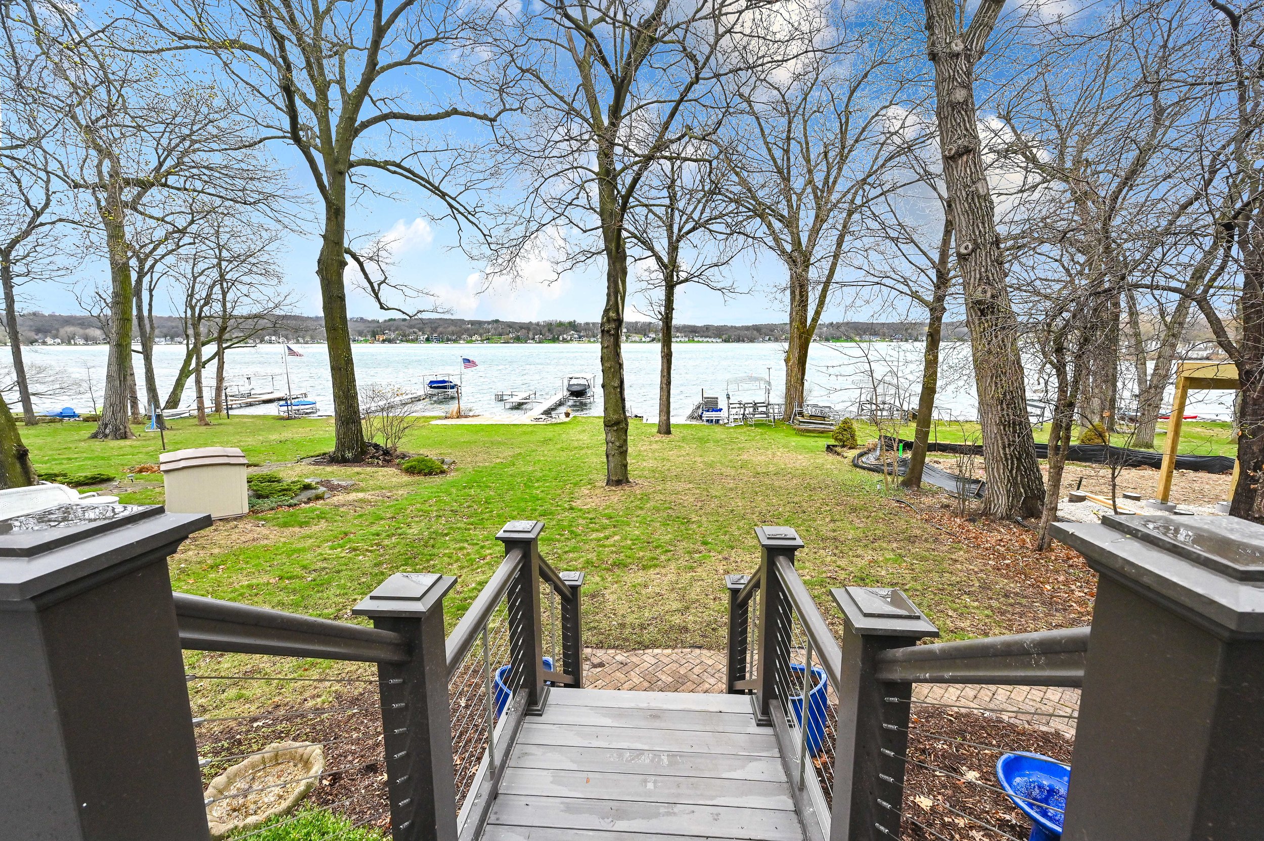 View from a deck looking out onto a grassy yard with trees and a lake with boats and docks in the background, under a partly cloudy sky.