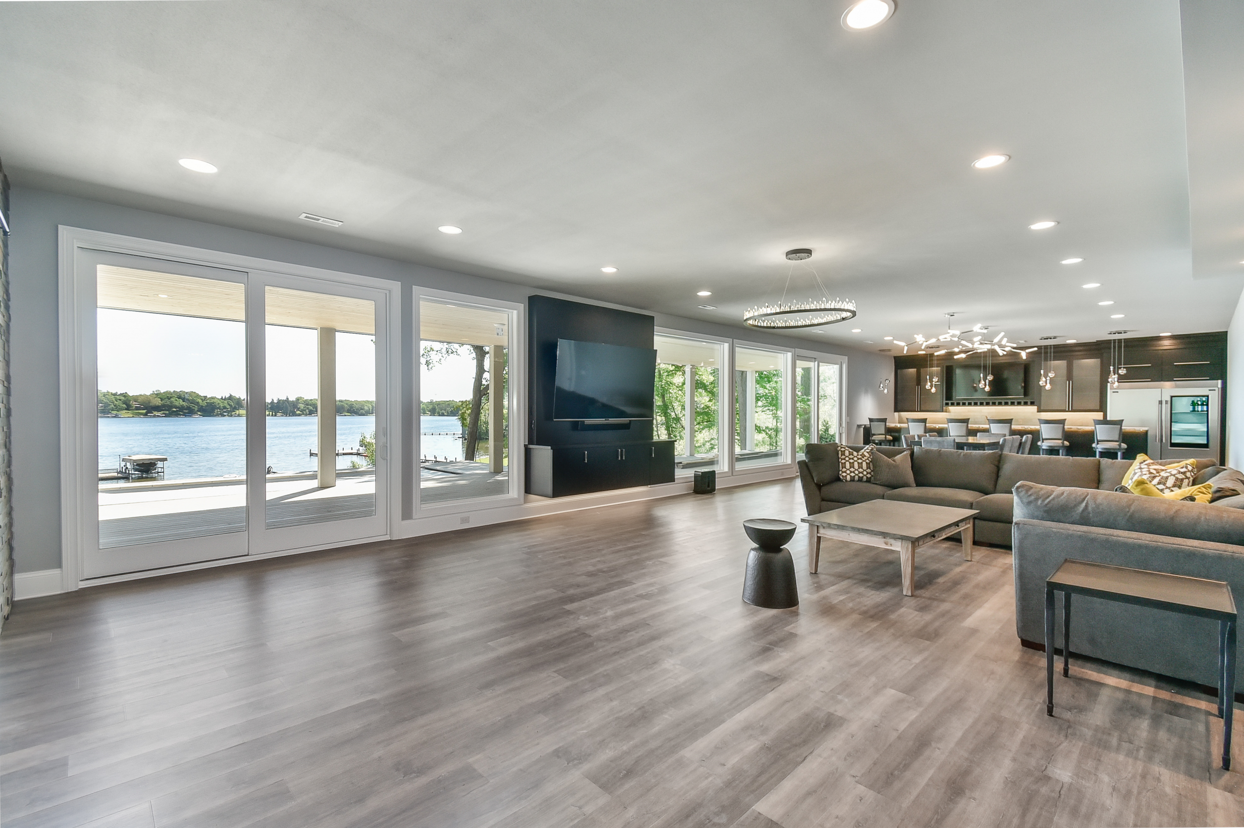 Spacious living room with large glass sliding doors overlooking a lake, featuring modern furnishings and a kitchen in the background.