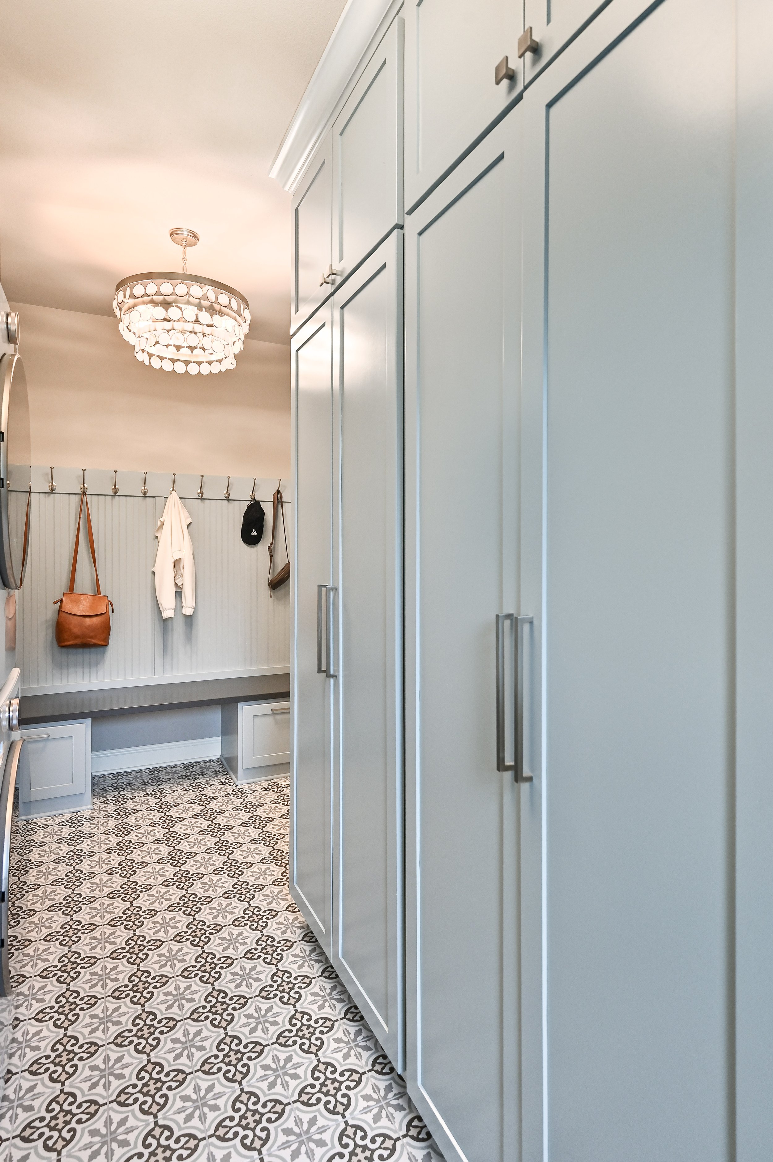 View of a laundry or mudroom with light blue cabinets, a patterned tile floor, and a wall with hooks holding bags and clothing, with a ceiling light fixture.