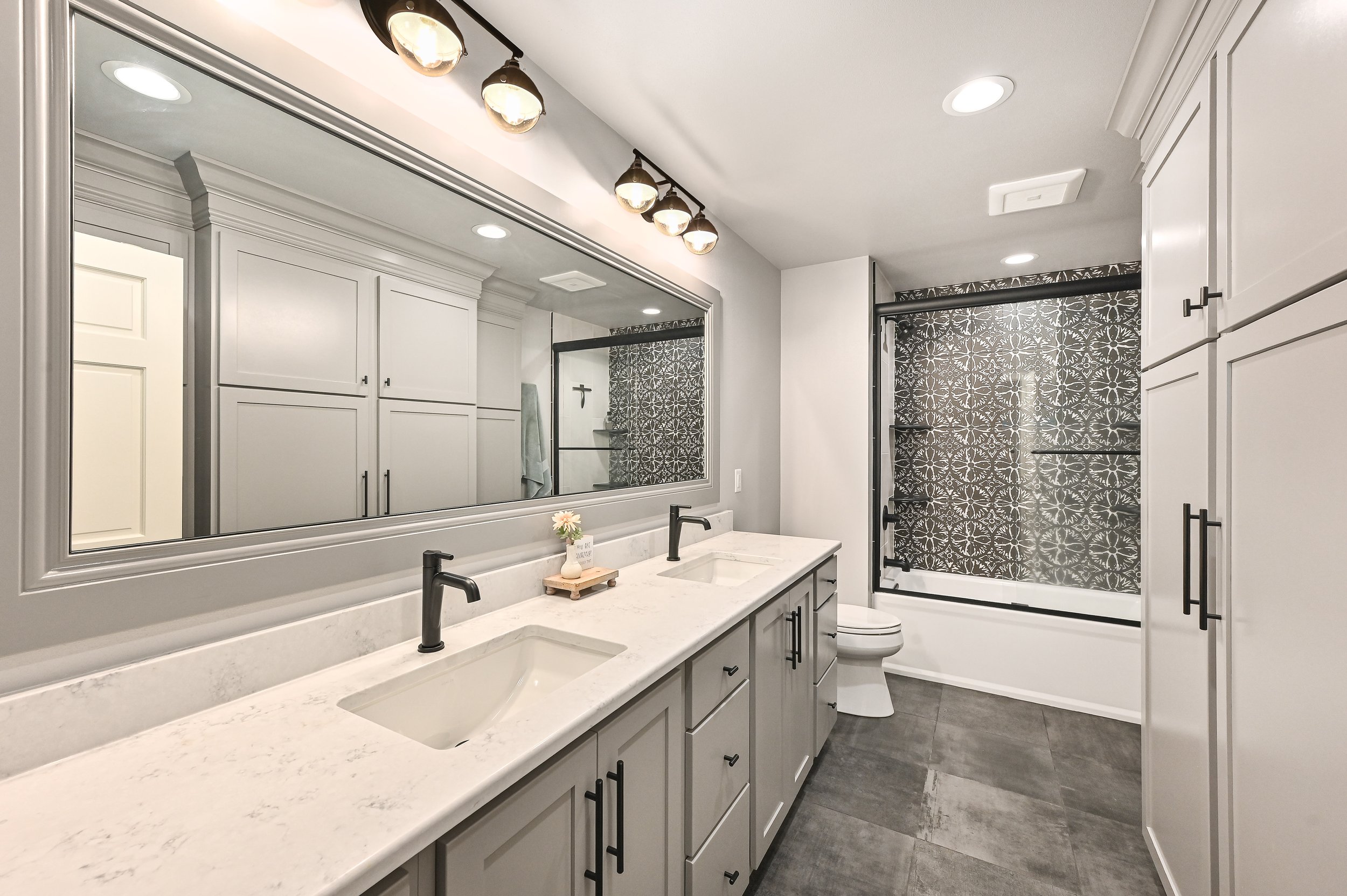 Modern bathroom with dual sinks, large mirror, gray cabinetry, black fixtures, a decorated shower with patterned tiles, and a separate toilet area.